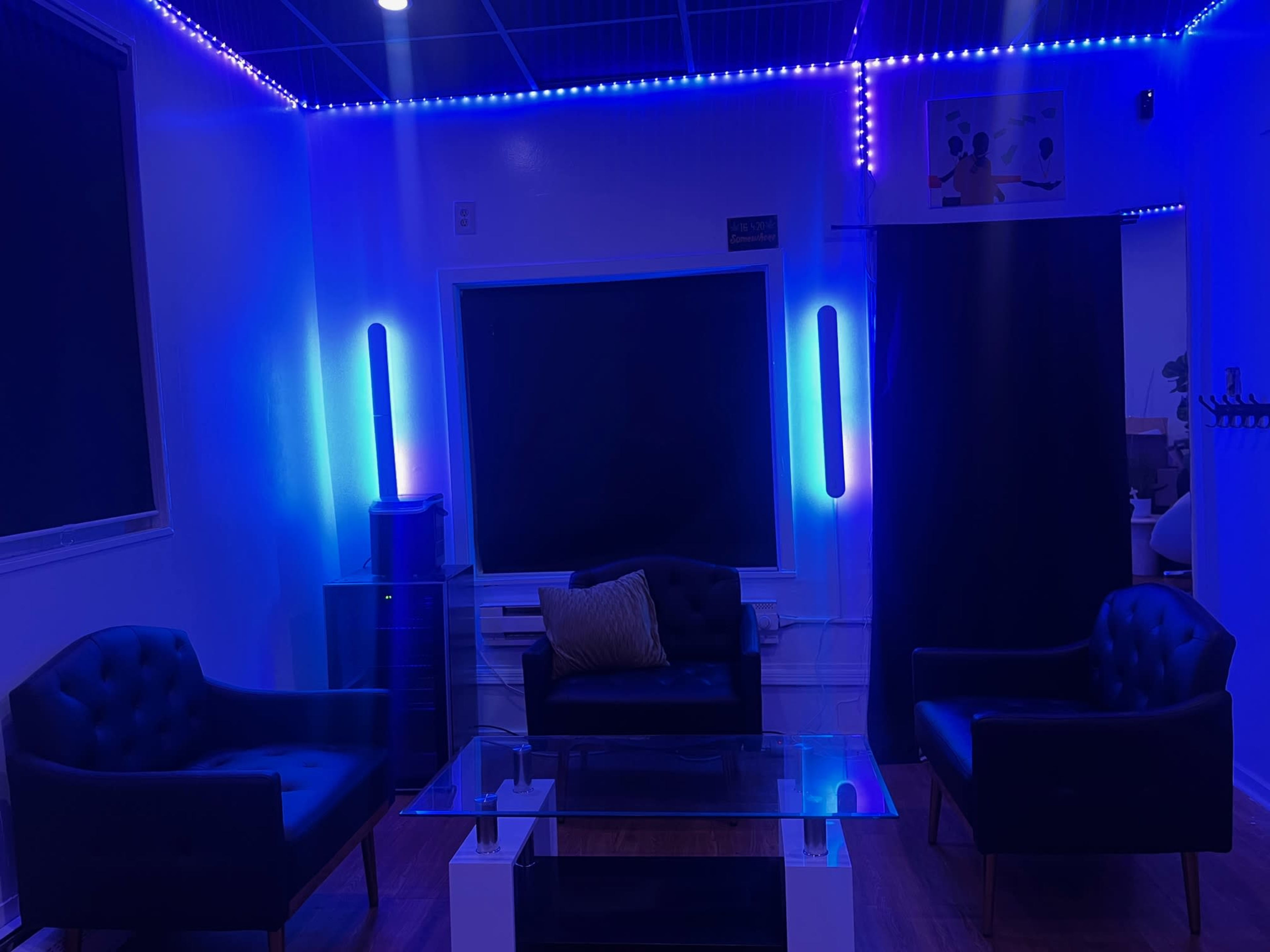 The image shows a room with two black chairs facing a glass table, a large screen on the wall, and ambient blue and purple lighting.