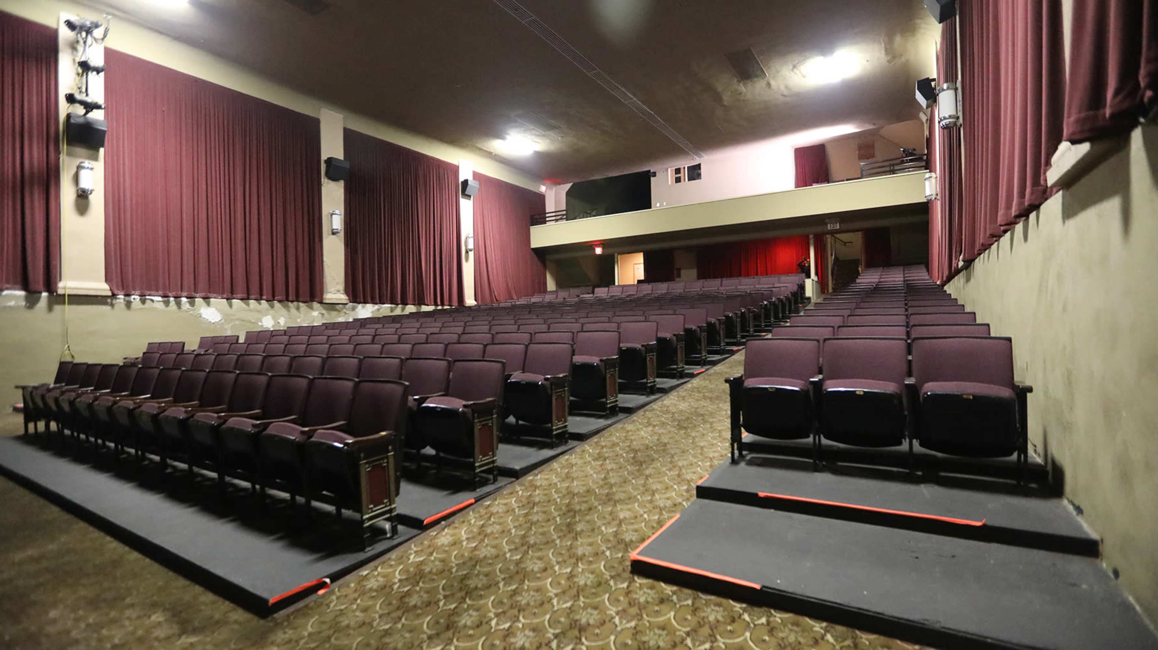 An interior view of a theater with empty rows of maroon seats and curtains lining the walls.