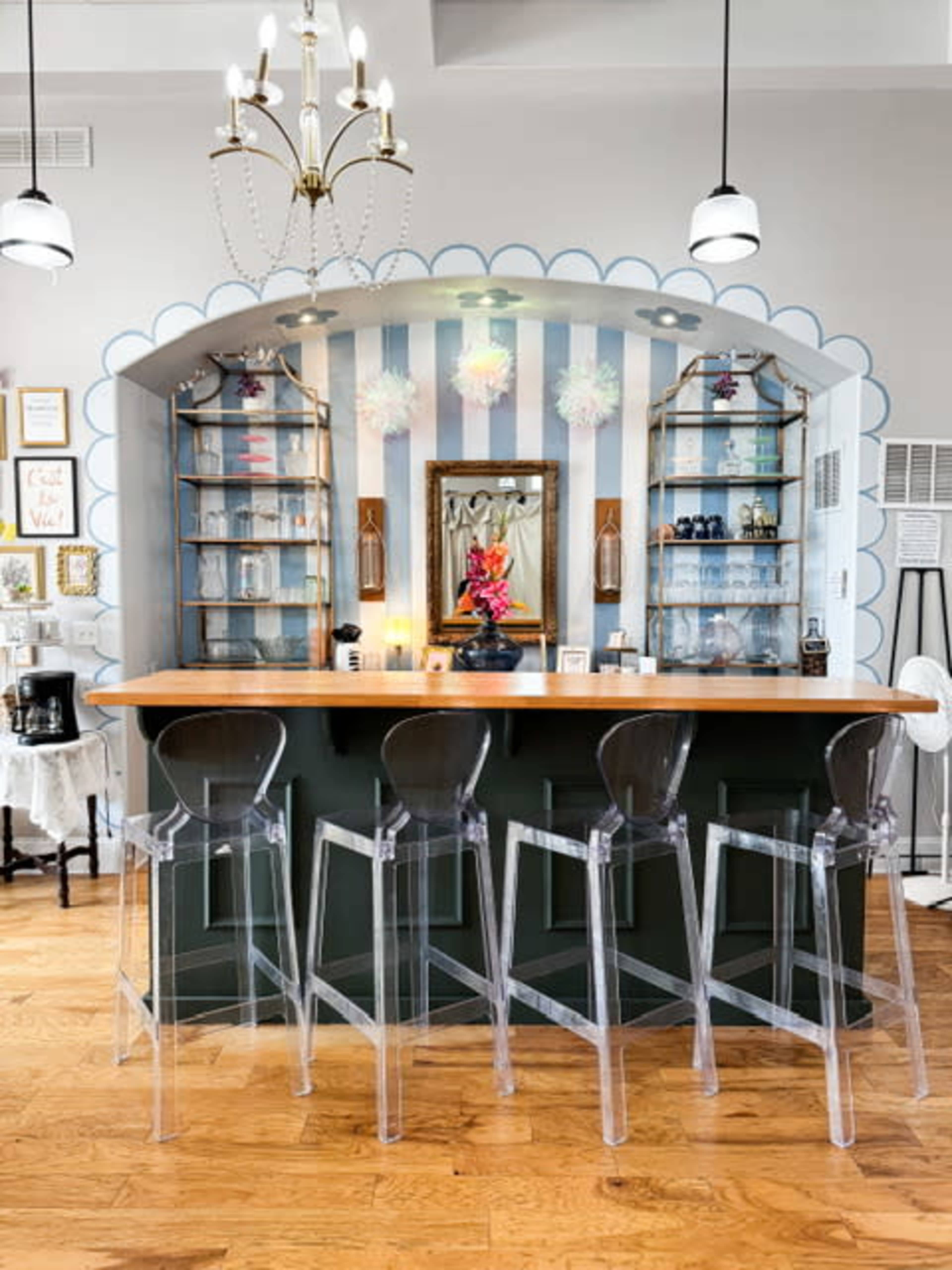 The image shows a stylish bar area with a wooden counter and transparent acrylic stools, set against a backdrop of striped walls and decorative shelves.