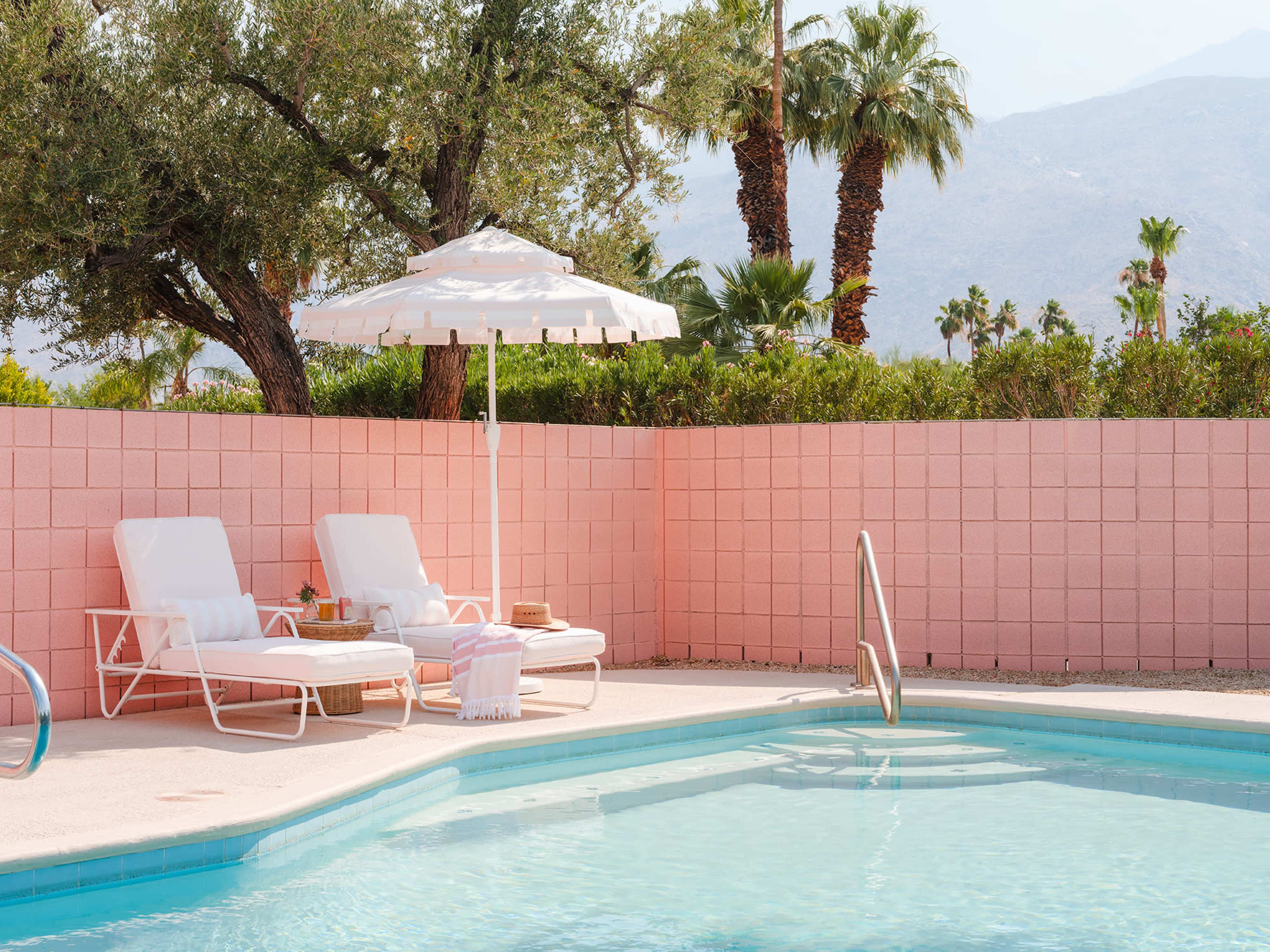 A pink-walled pool area with two lounge chairs, an umbrella, and palm trees in the background.