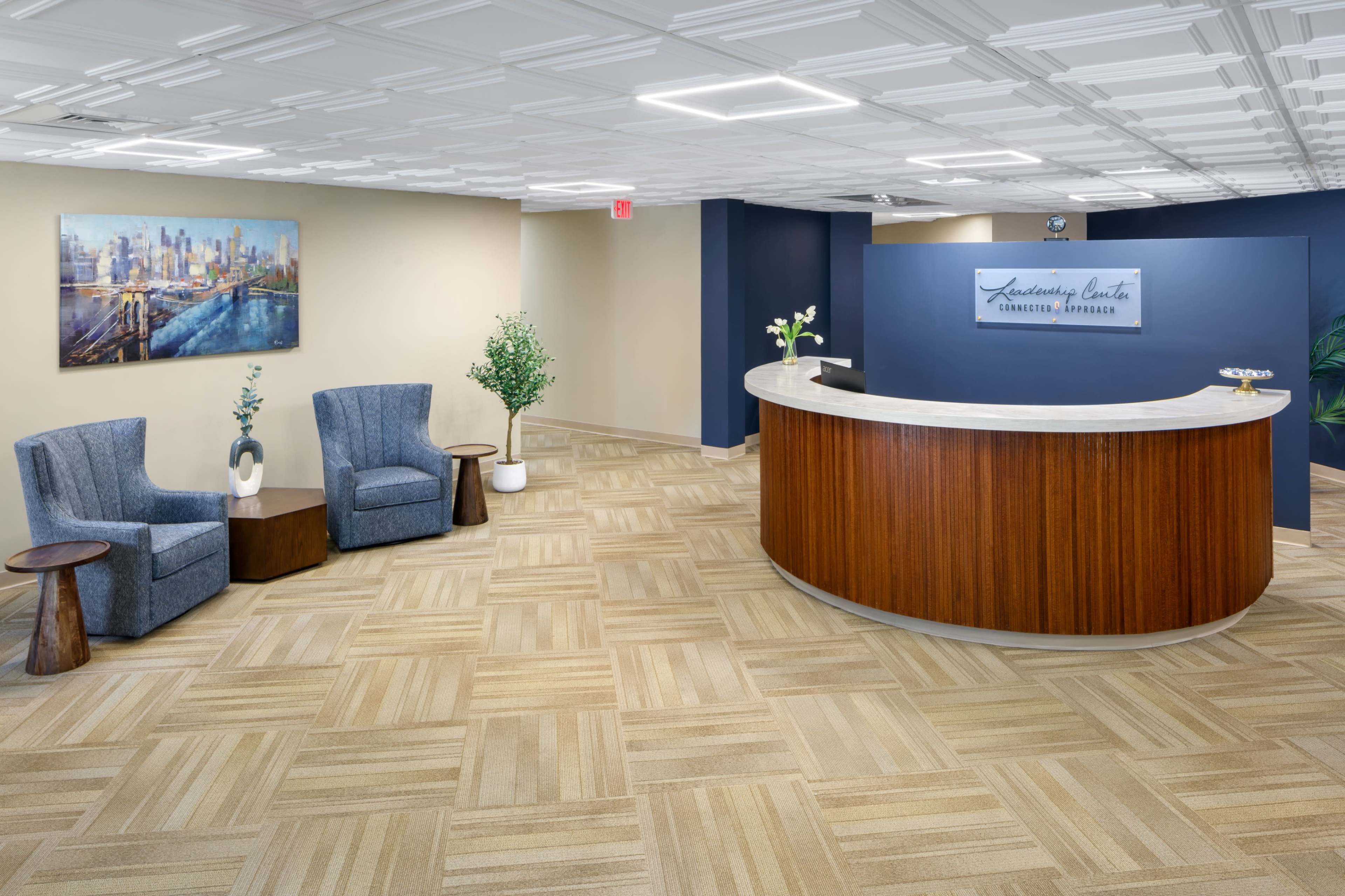 The image shows a modern reception area with two gray chairs, a round wooden table, and a curved front desk with a sign, set against a light-colored wall and stylish flooring.