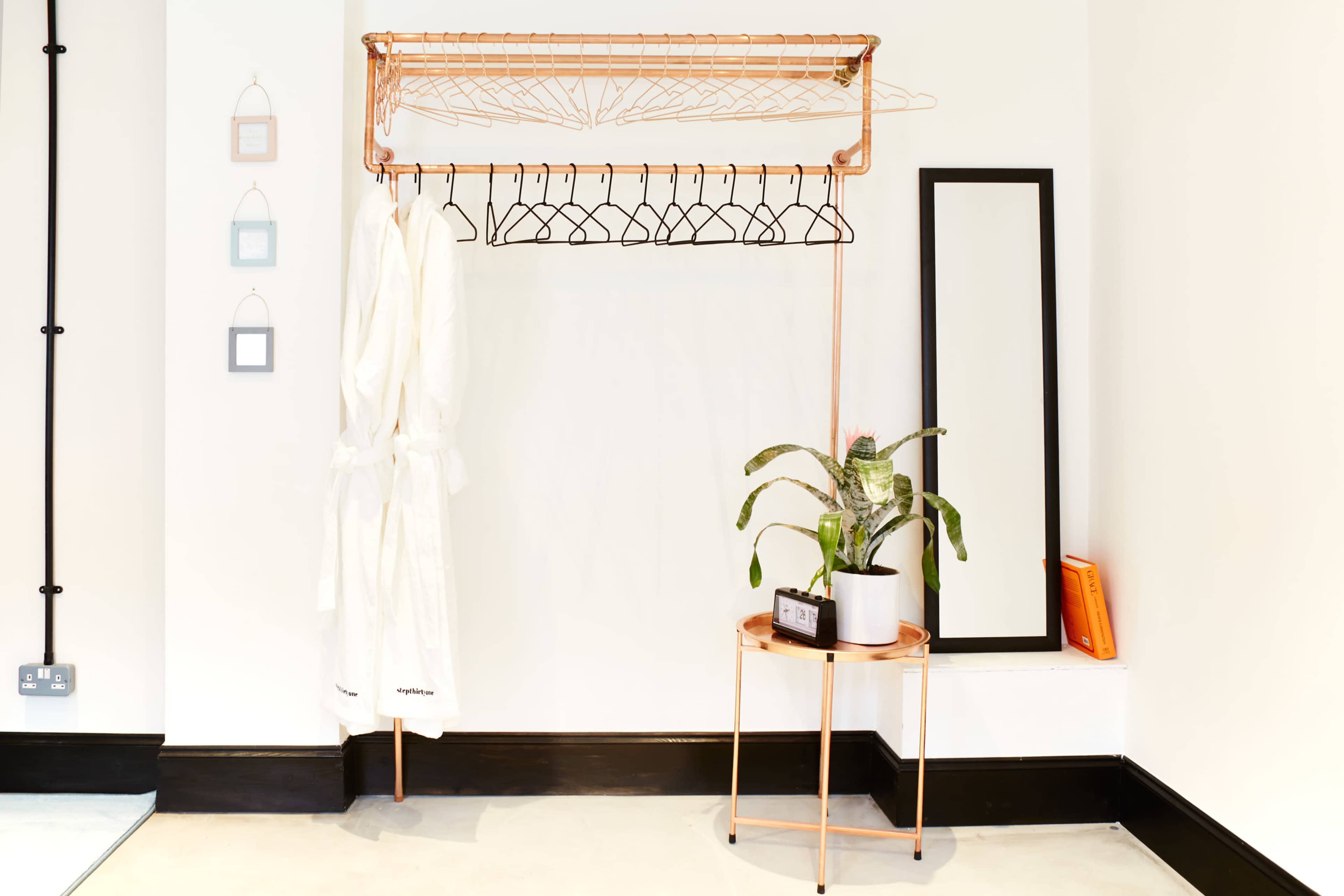 A copper clothing rack holds several hangers and a white robe, next to a mirror and a potted plant on a small table against a white wall.