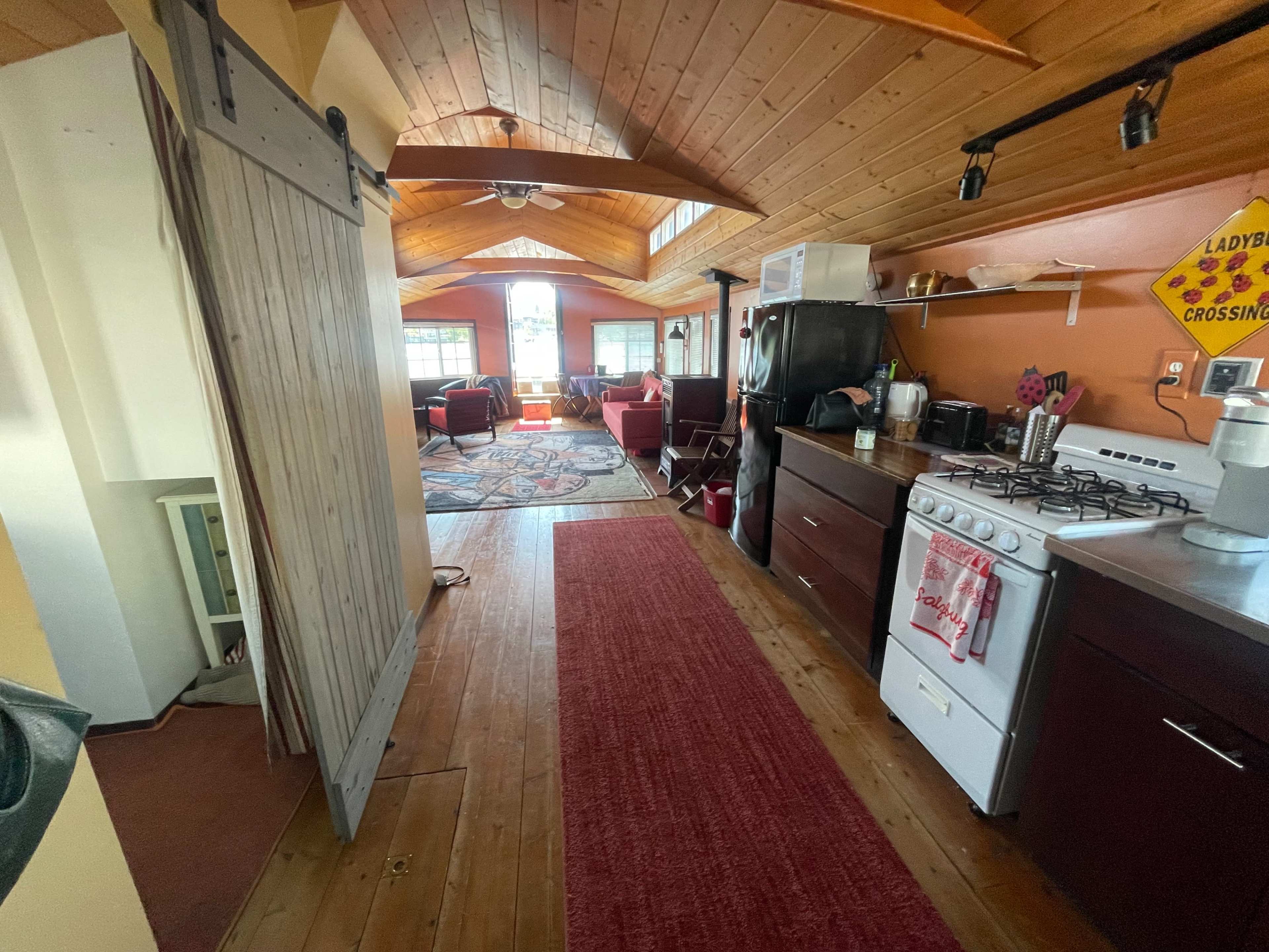 The image shows a cozy interior space with a wooden ceiling, an open kitchen area, and a seating section filled with chairs and a rug.