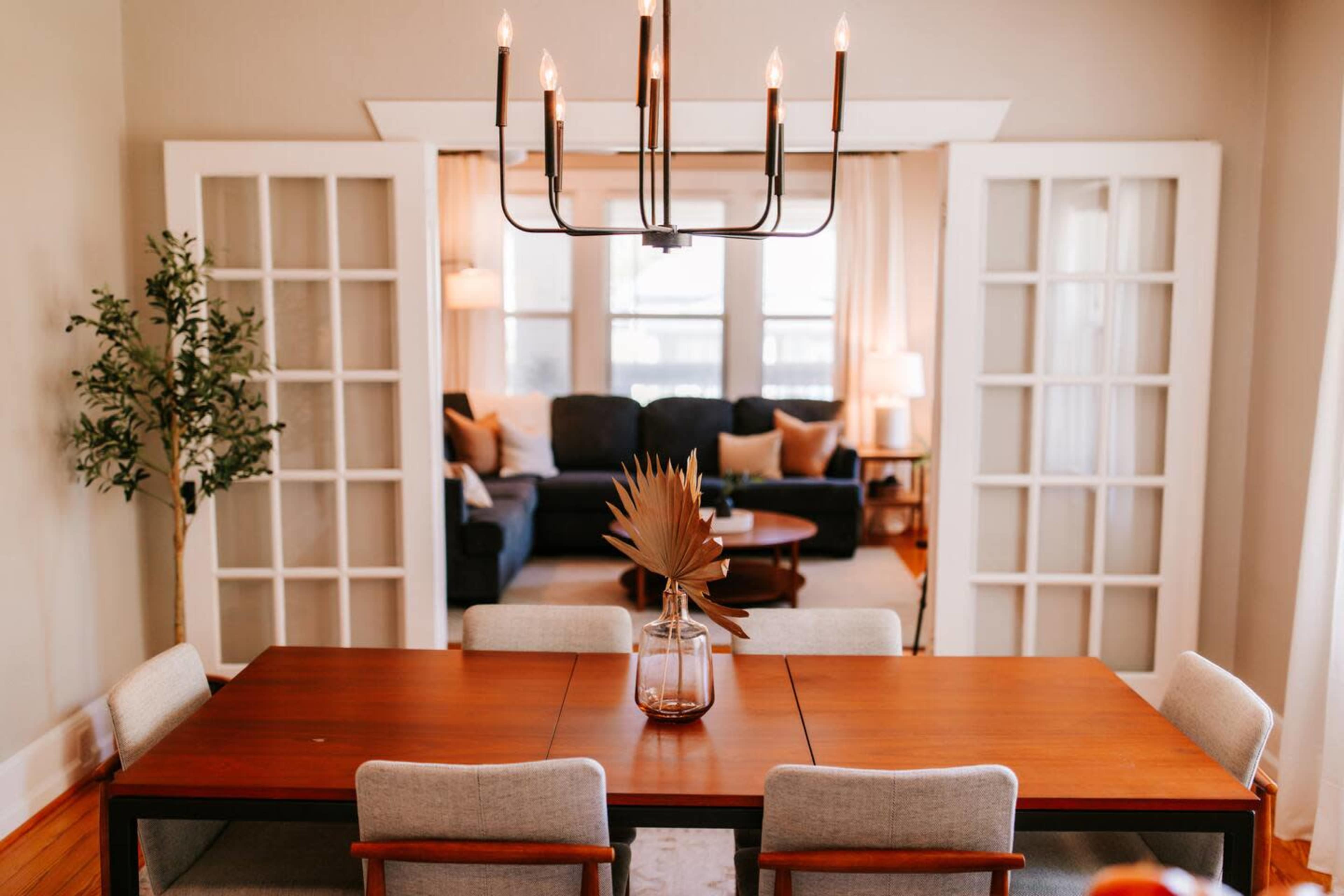 The image shows a dining area with a wooden table and chairs, a chandelier above, and a living space visible in the background through double doors.