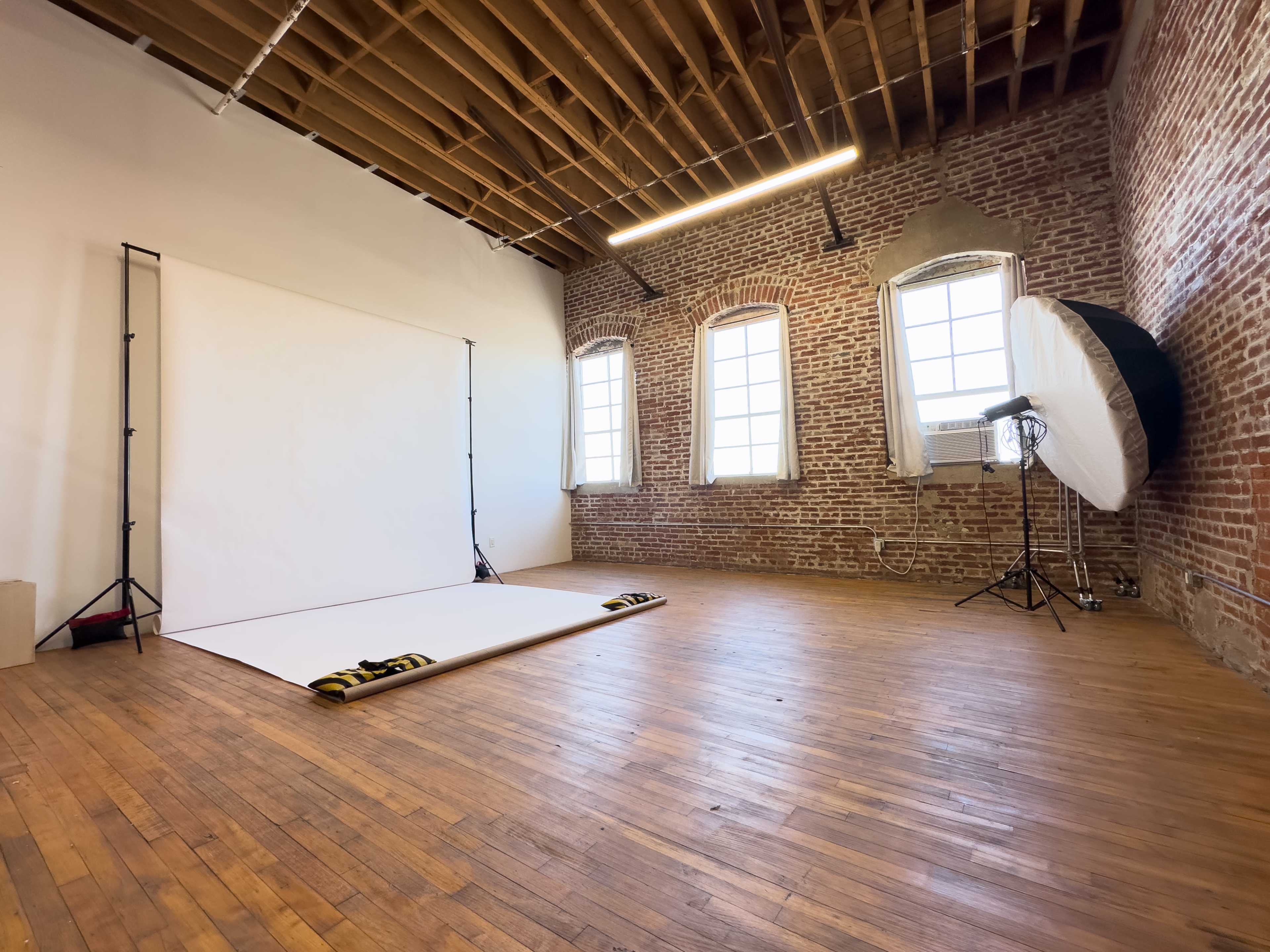 The image shows a spacious photography studio with a white backdrop, large windows, exposed brick walls, and wooden flooring.
