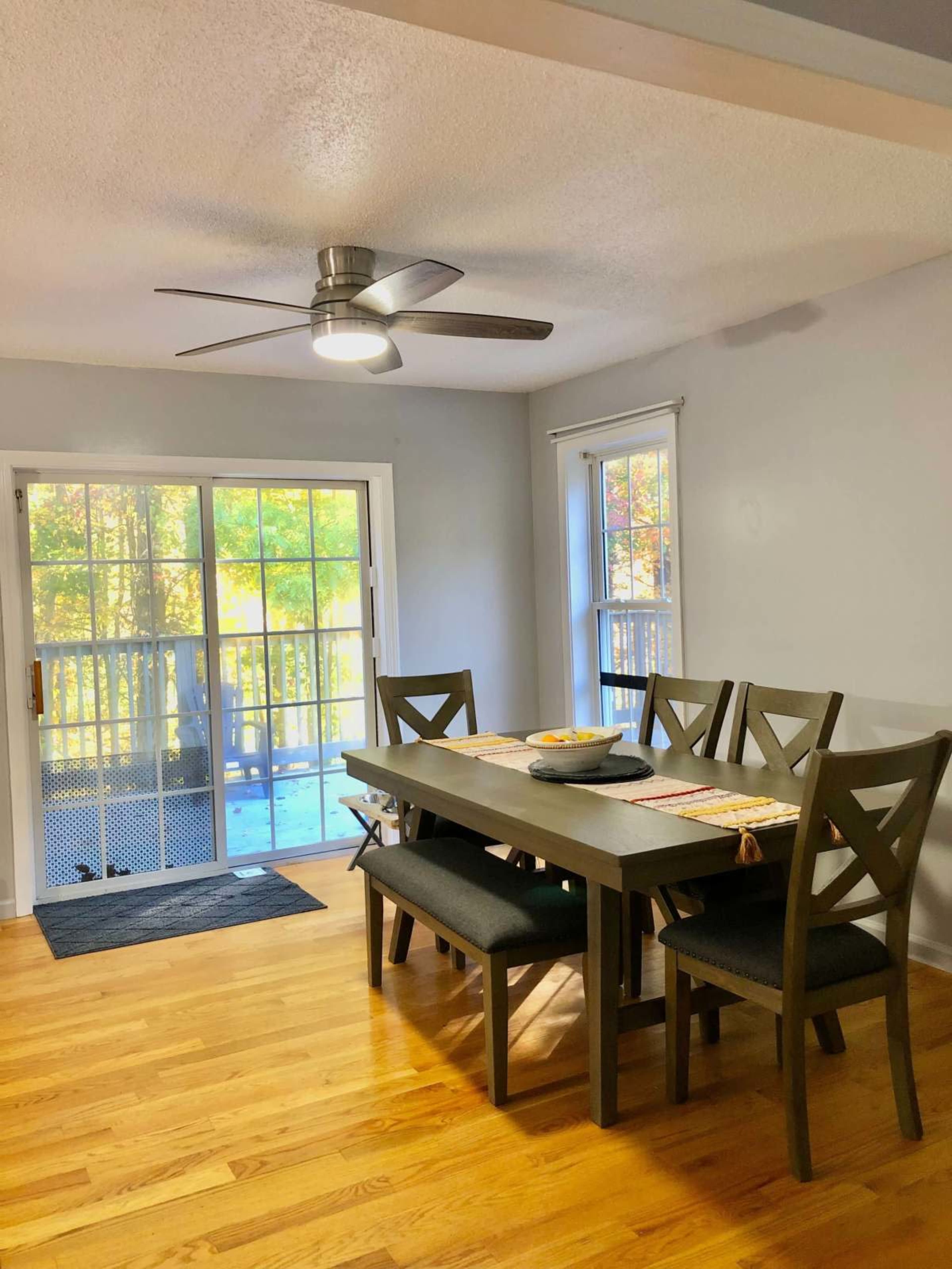 The image shows a dining area with a wooden table and chairs, a ceiling fan, and a door leading to a deck outside.