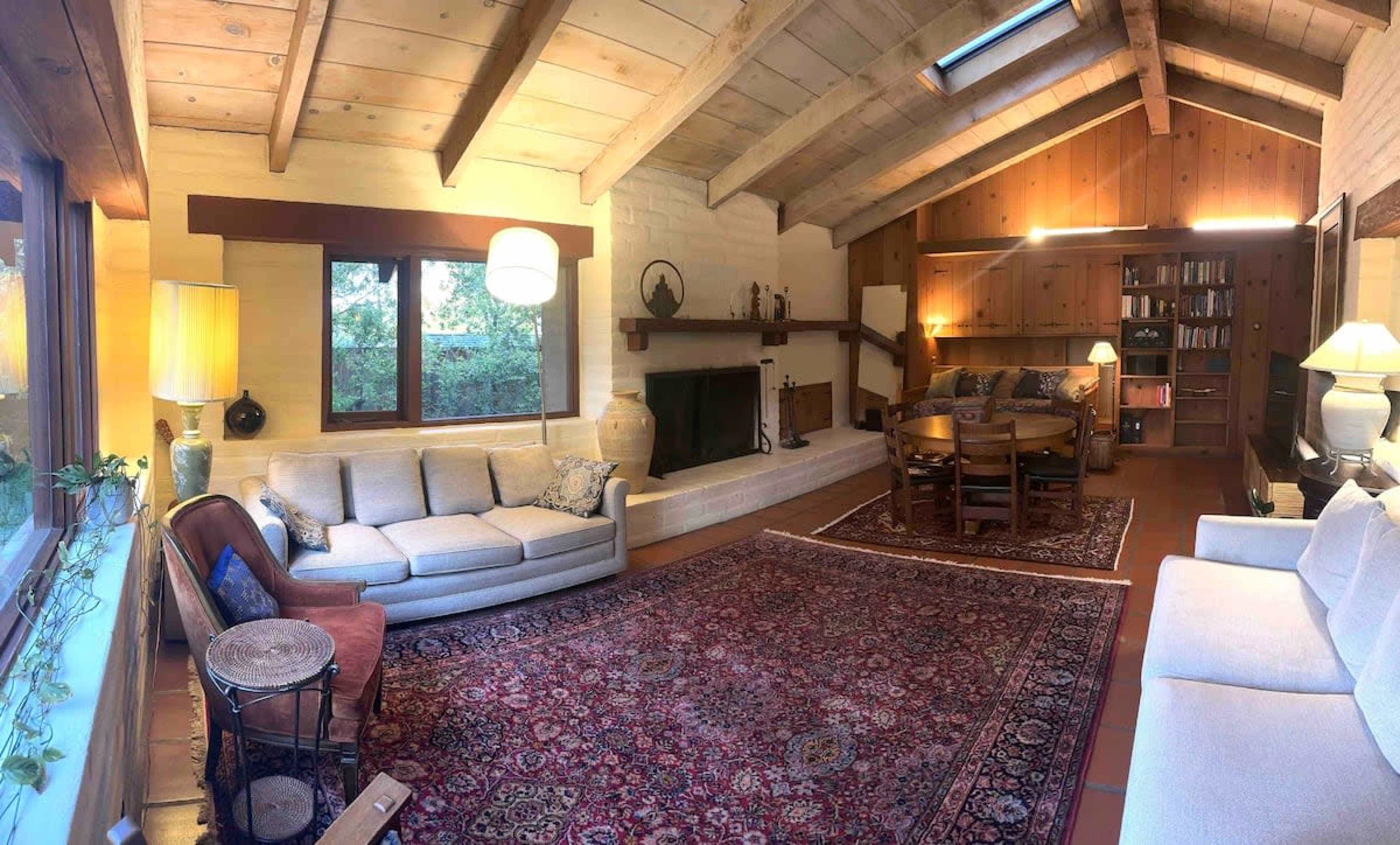 A spacious living room featuring a central area rug, a wooden ceiling, a fireplace, and seating arrangements including a sofa and dining table.