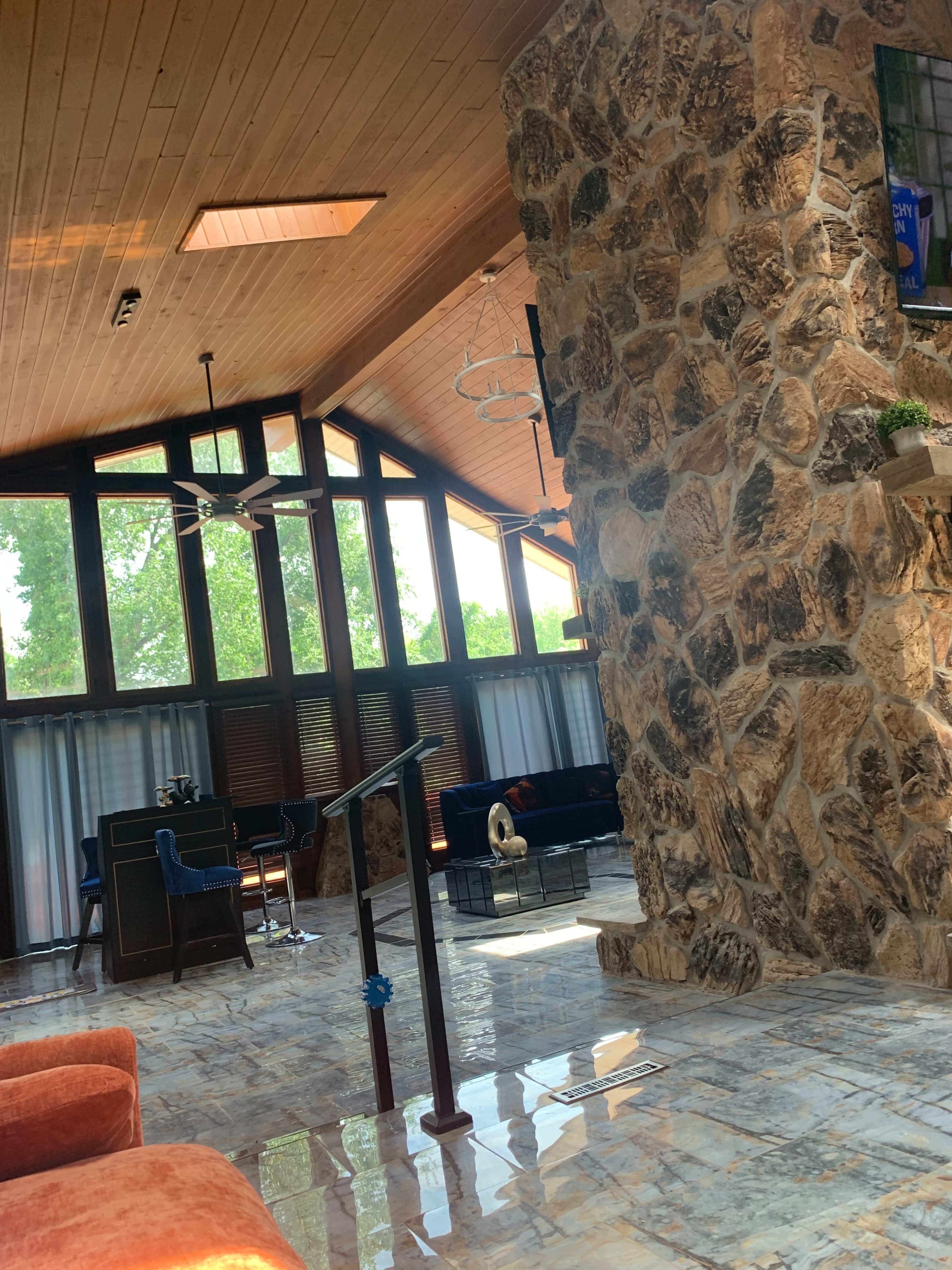 The image shows a spacious living area with a high wood-paneled ceiling, large windows, a stone wall, and modern furniture.