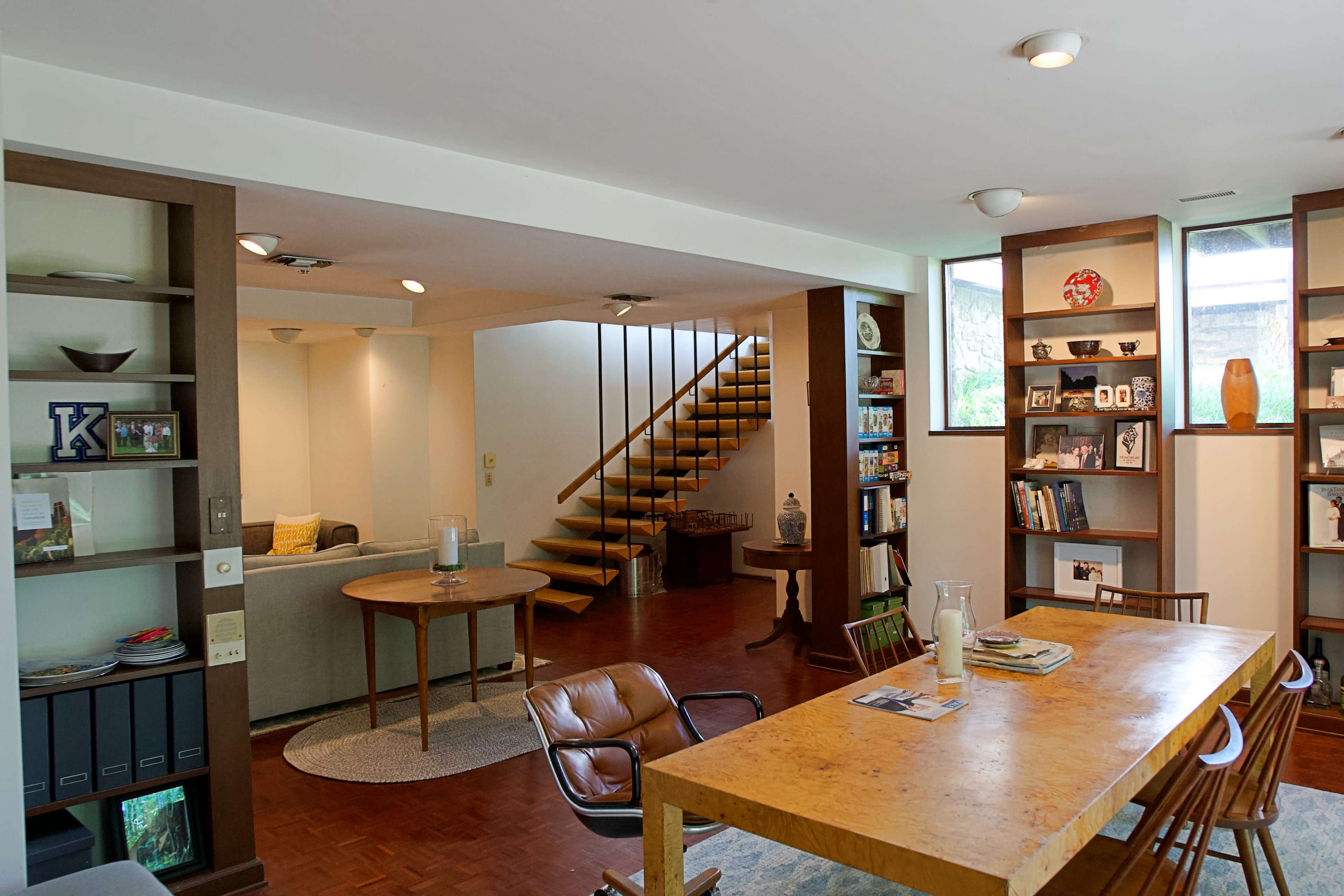 The image shows a modern living space with a dining table, wooden chairs, bookshelves, and a staircase leading to an upper floor.