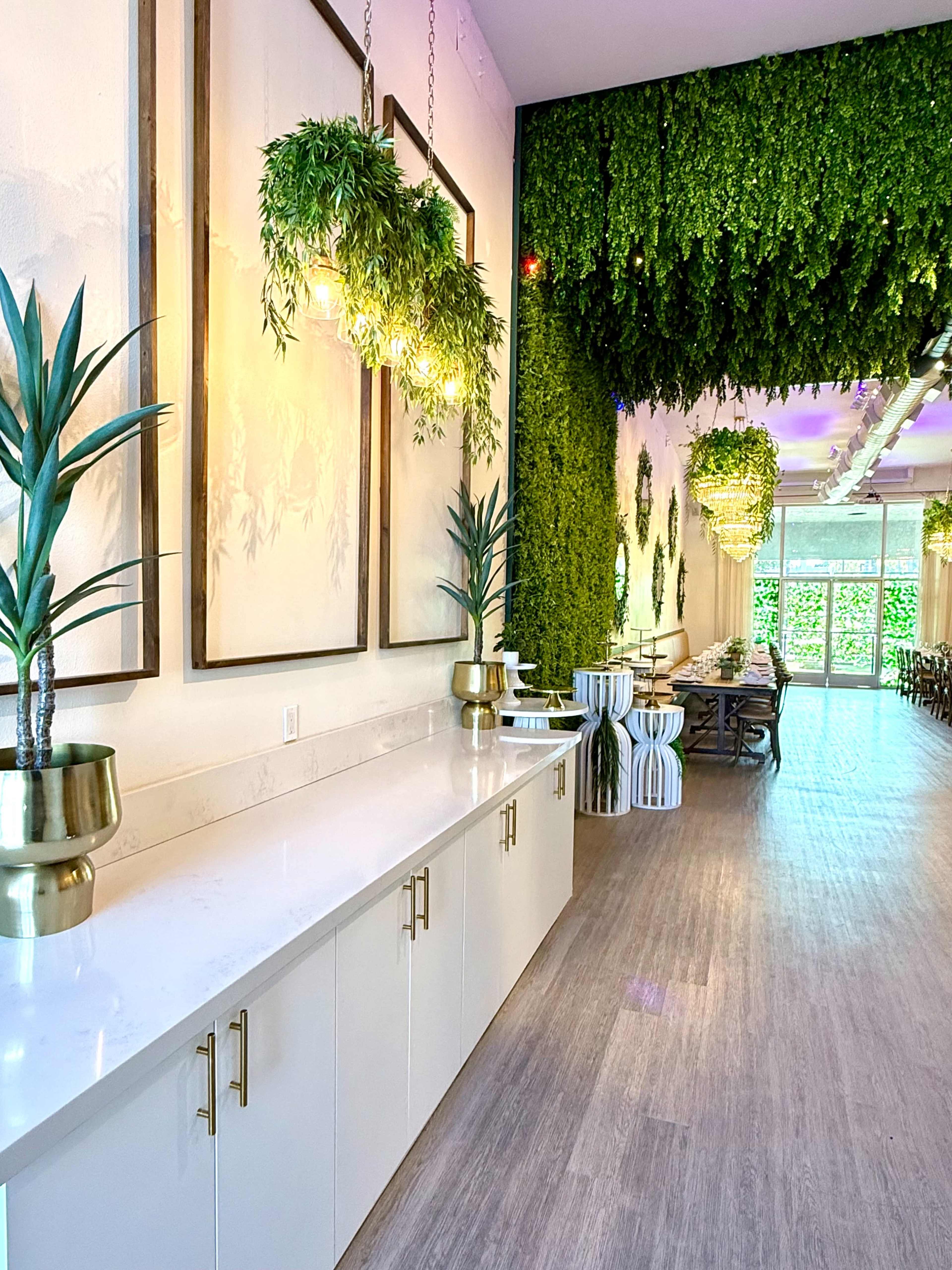 The image shows a modern interior space featuring a long, narrow hallway with light-colored cabinetry, potted plants, and illuminated fixtures, accented by lush greenery along the walls.