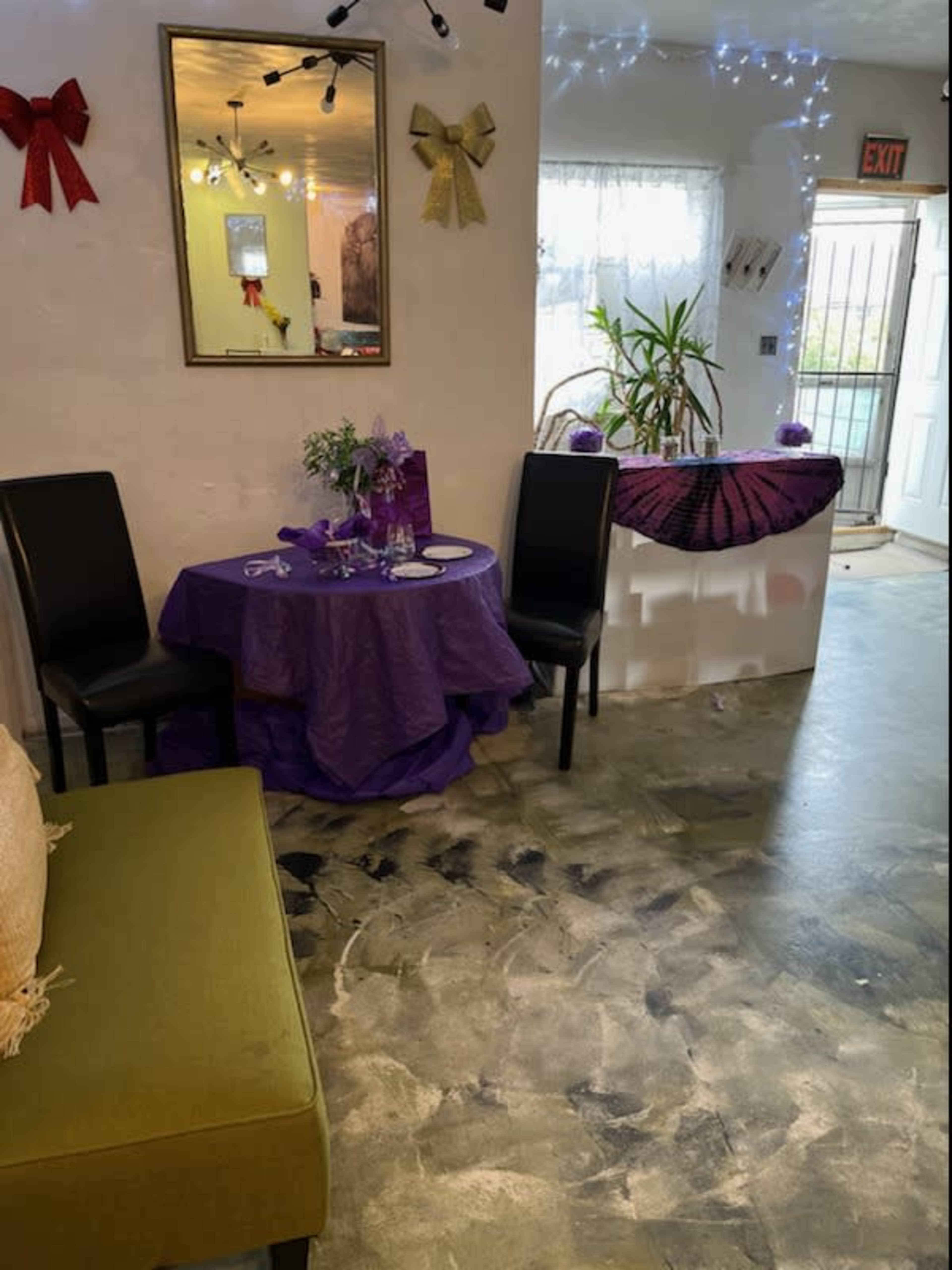 The image shows a small, decorated room with a purple tablecloth on a round table, two black chairs, and a plant near the entrance.