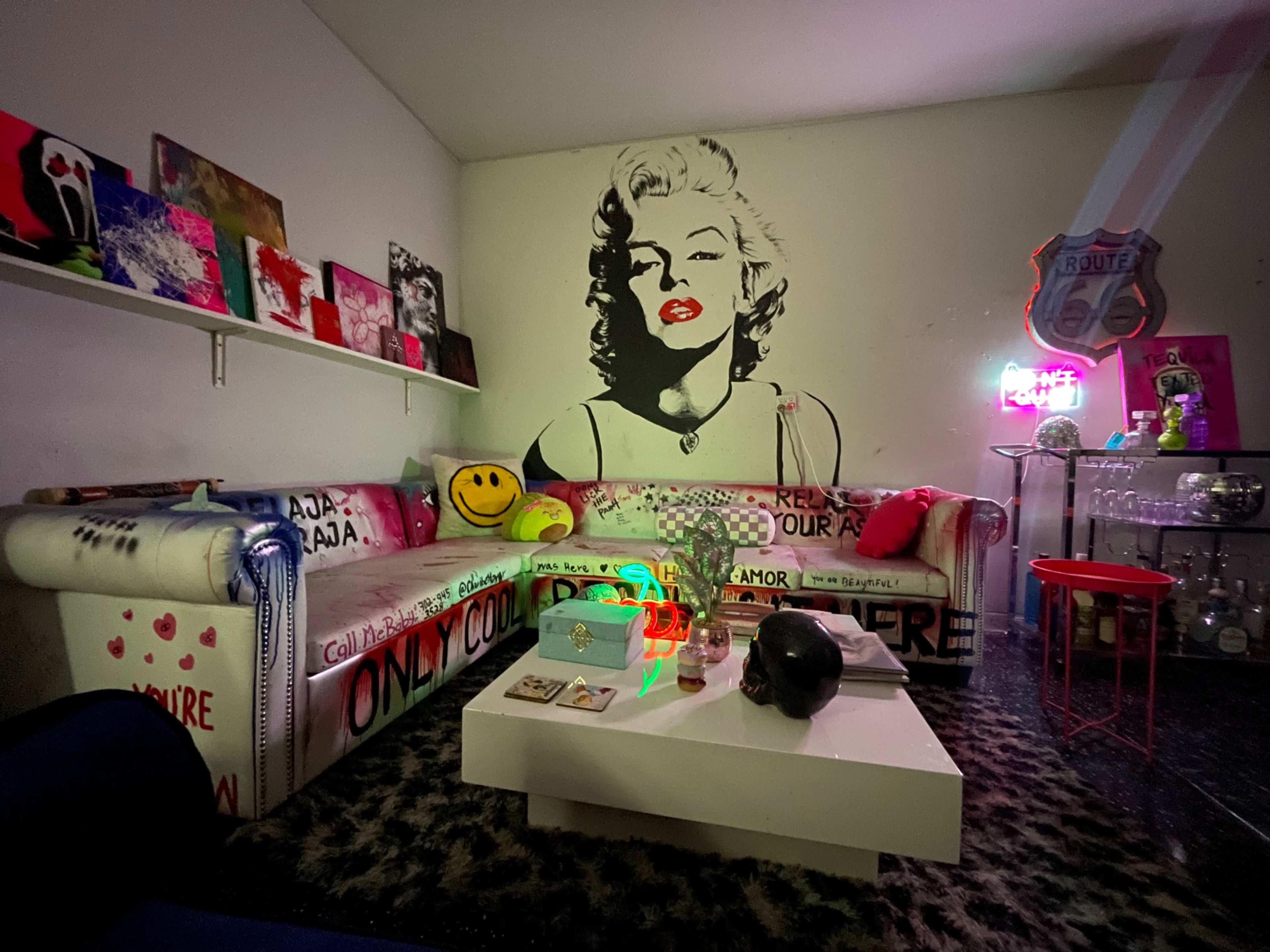 The image shows a vibrant living room featuring a mural of Marilyn Monroe on the wall, a colorful couch with decorative pillows, and a coffee table adorned with various items.