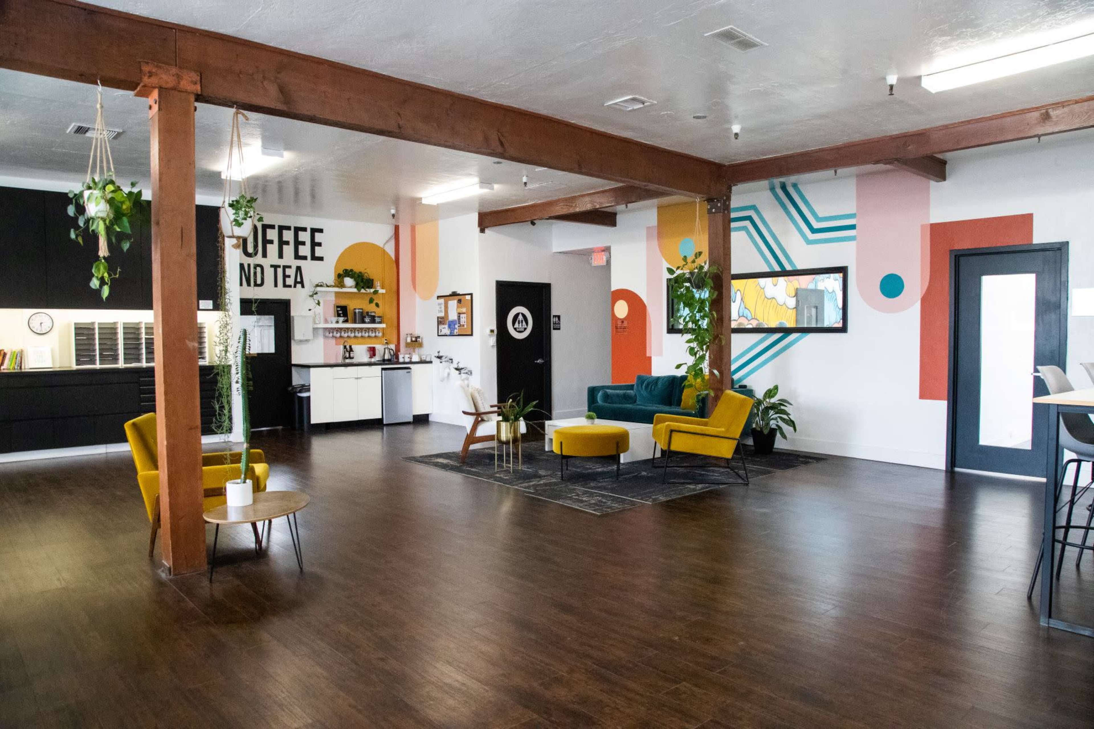 The image shows a spacious, modern interior with wooden beams, colorful wall designs, a seating area featuring yellow chairs, and a coffee and tea station.