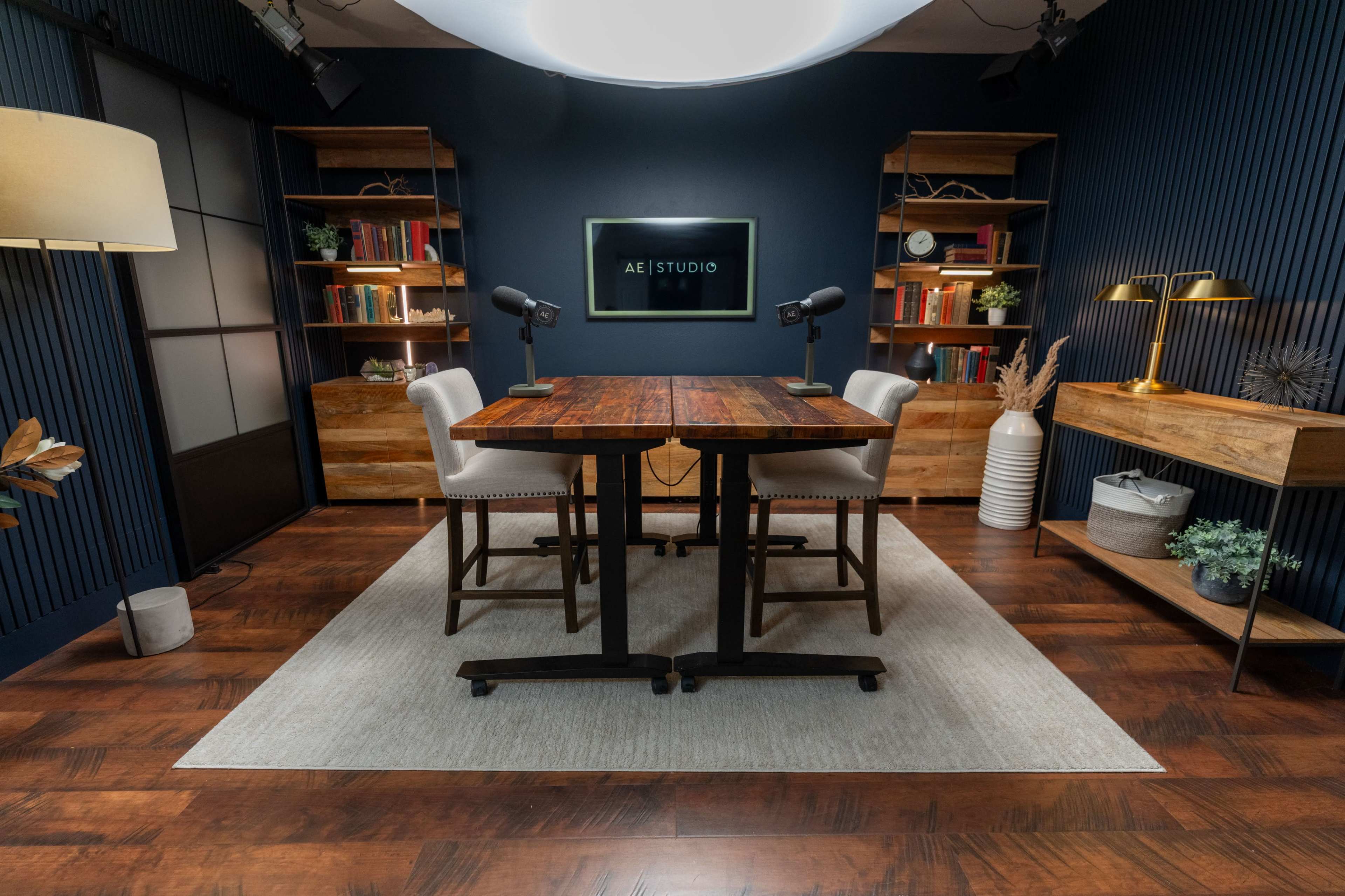 The image shows a modern studio setup featuring a wooden table with four chairs, framed by dark blue walls and shelves lined with books and decorative items.