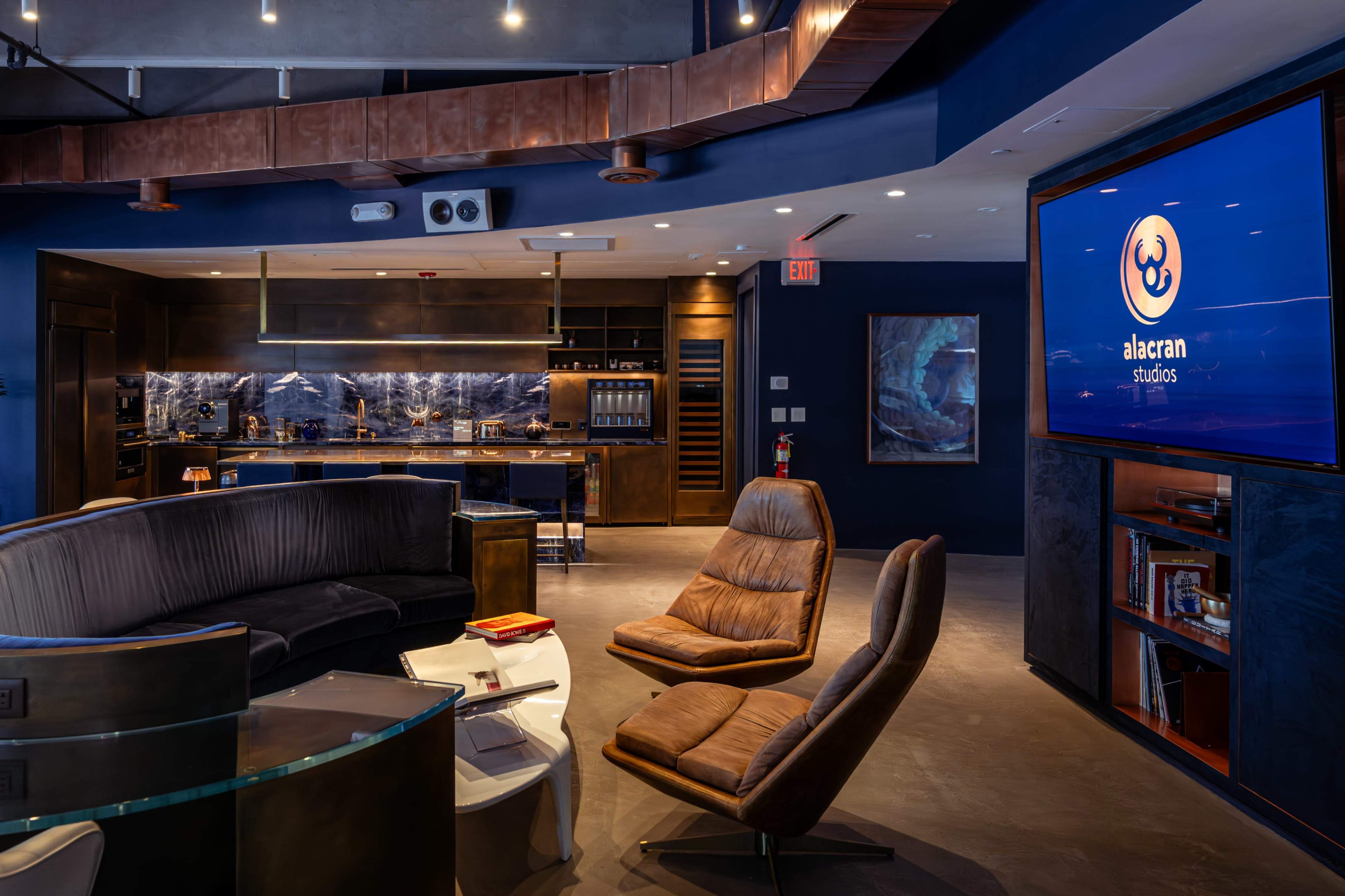 The image shows a modern studio interior featuring a curved couch, leather armchairs, and a large screen displaying the logo for "Alacran Studios," alongside a sleek kitchen area.