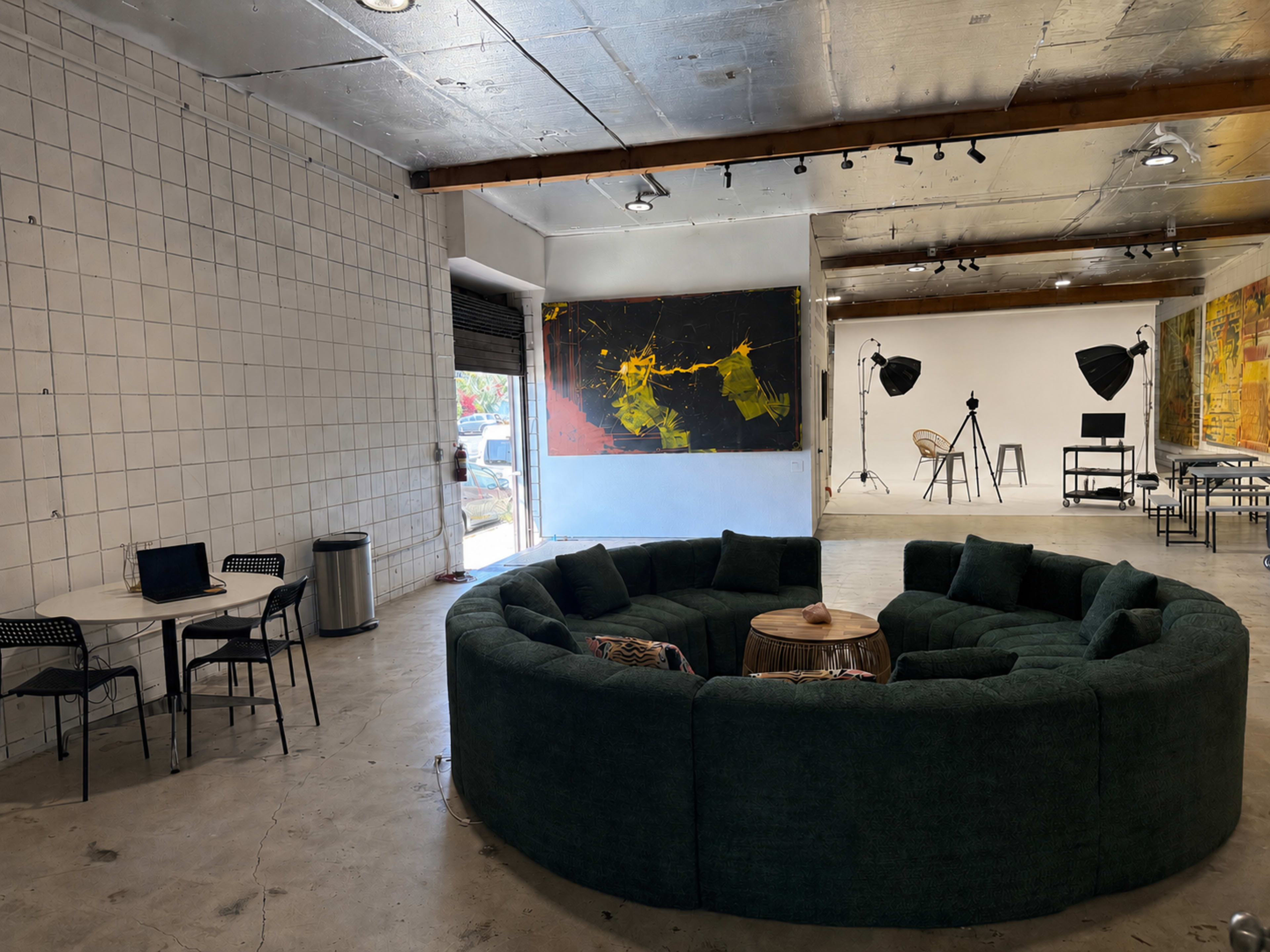 The image shows a spacious interior with a large, circular green sofa, a wooden coffee table in the center, and a dining area with a small table and chairs, while art pieces and photography equipment are visible in the background.