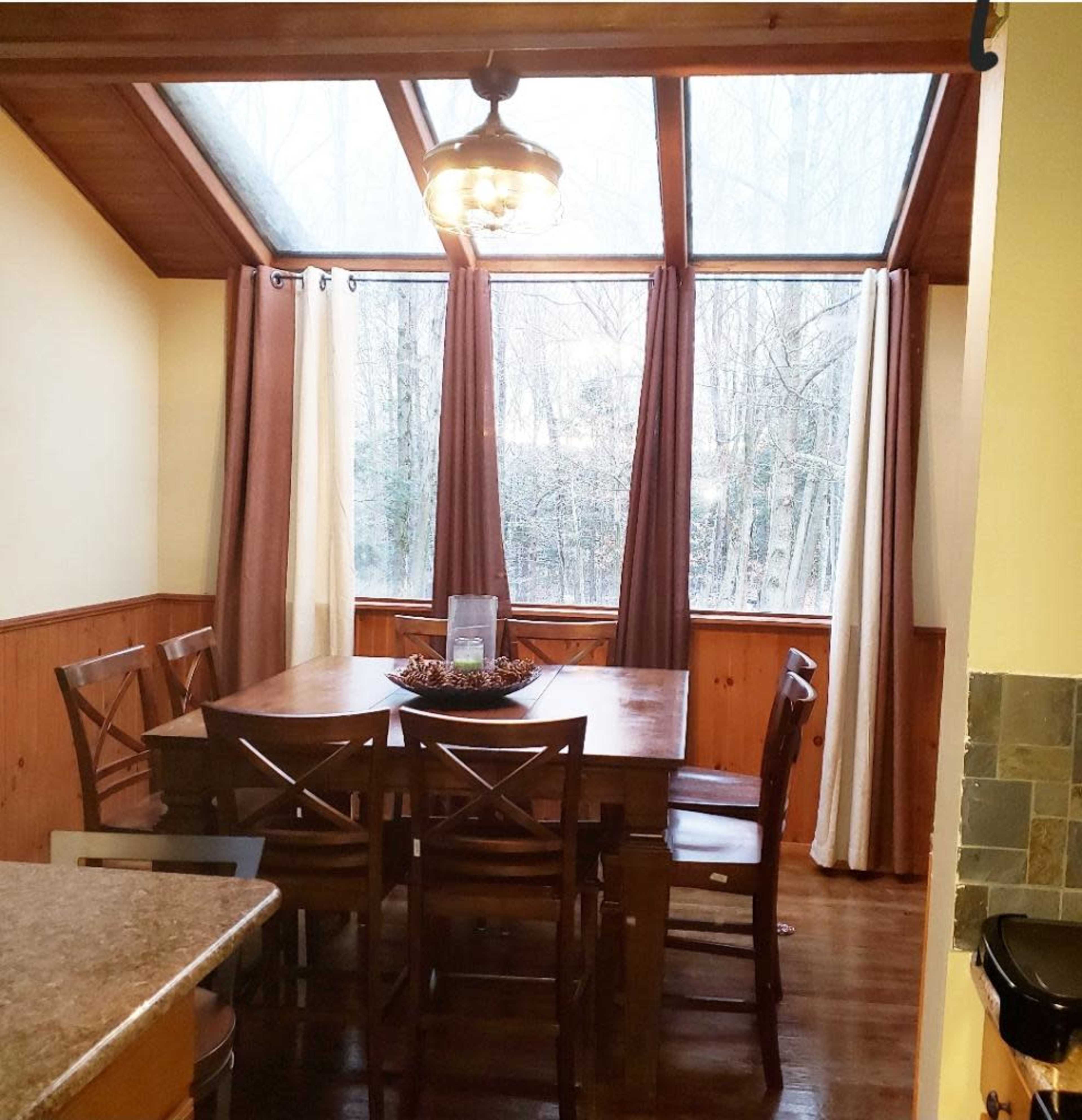 A wooden dining table surrounded by chairs is positioned in a sunlit room with large windows and curtains.