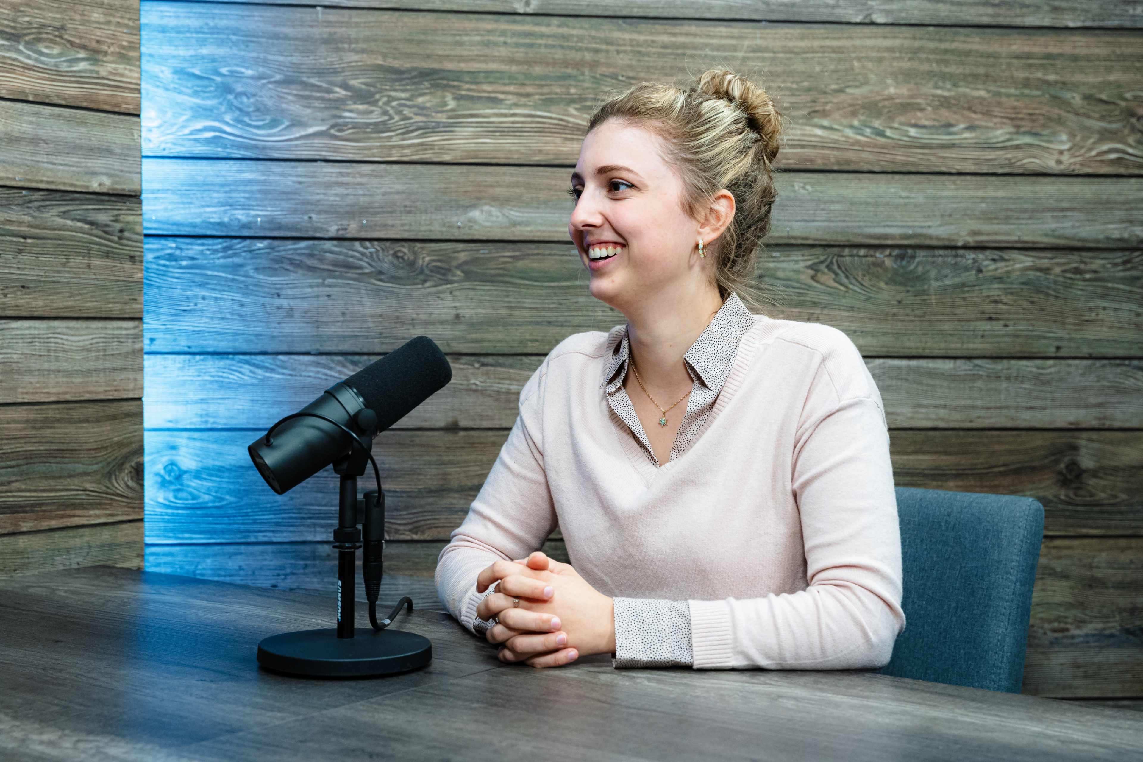 A woman is sitting at a table with a microphone in front of her, smiling while engaging in a conversation.