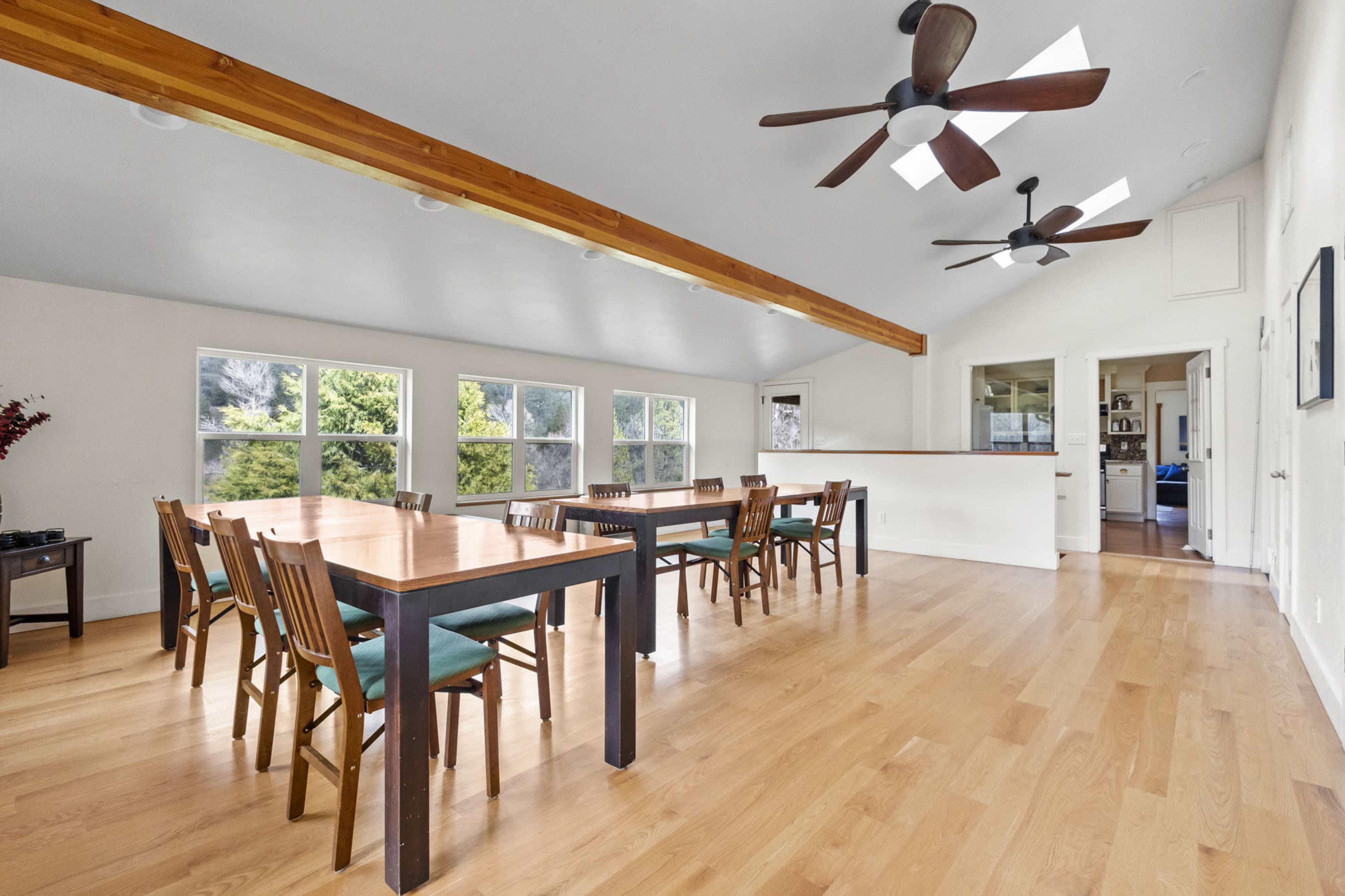 A spacious dining area features a long table and multiple chairs, illuminated by natural light from large windows.