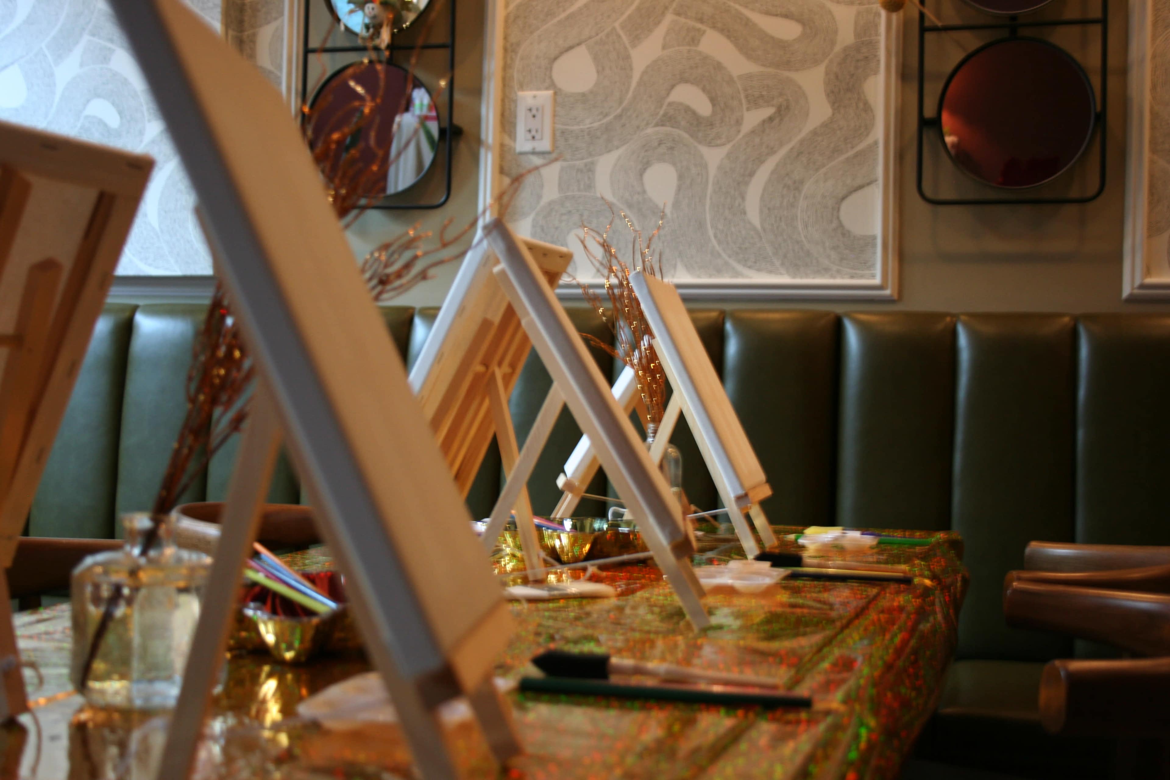The image shows a row of empty easels set up on a table adorned with colorful art supplies and decorative elements in a well-lit interior space.