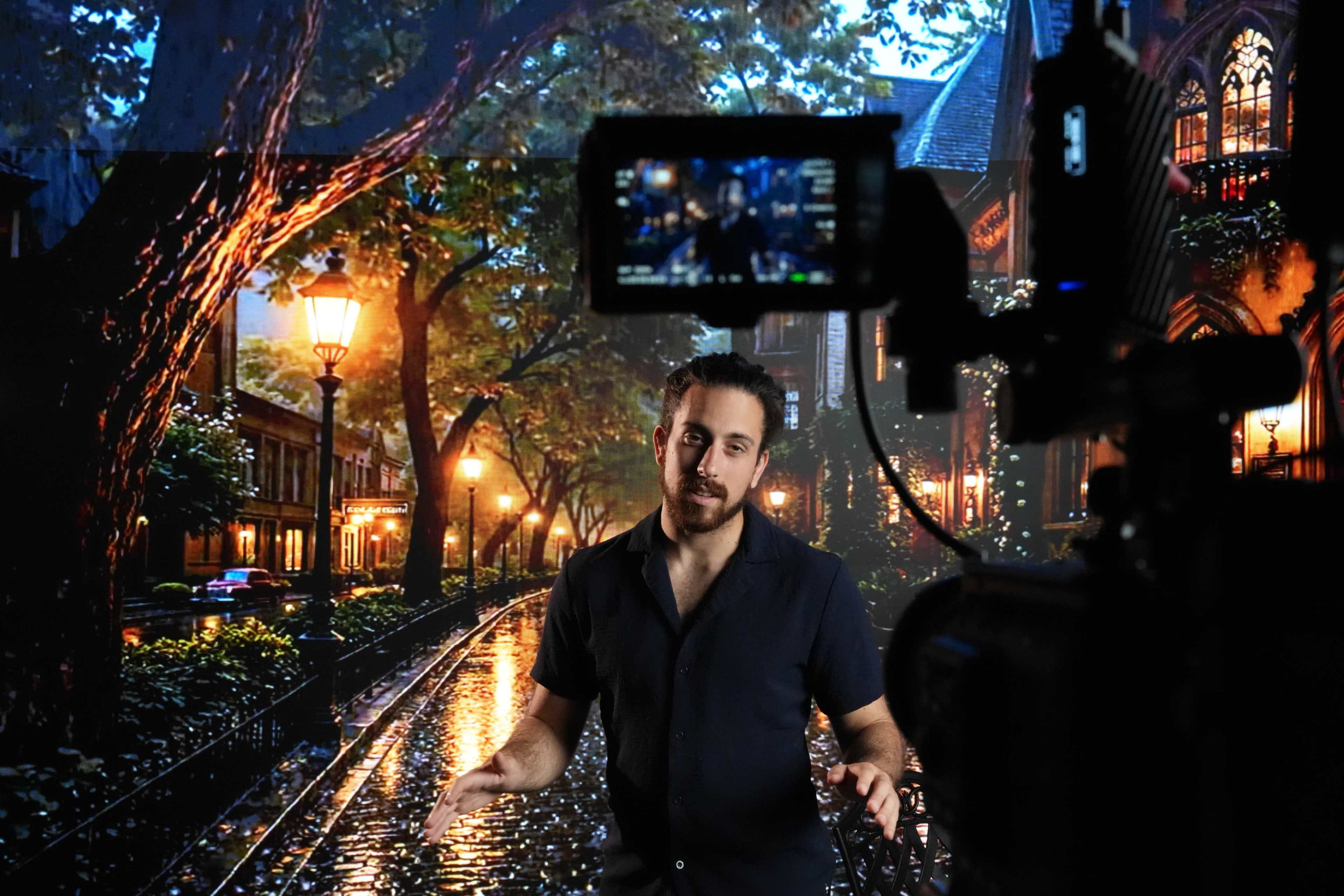 A man stands in front of a camera, speaking against a backdrop of a lit street scene with trees and wet pavement.
