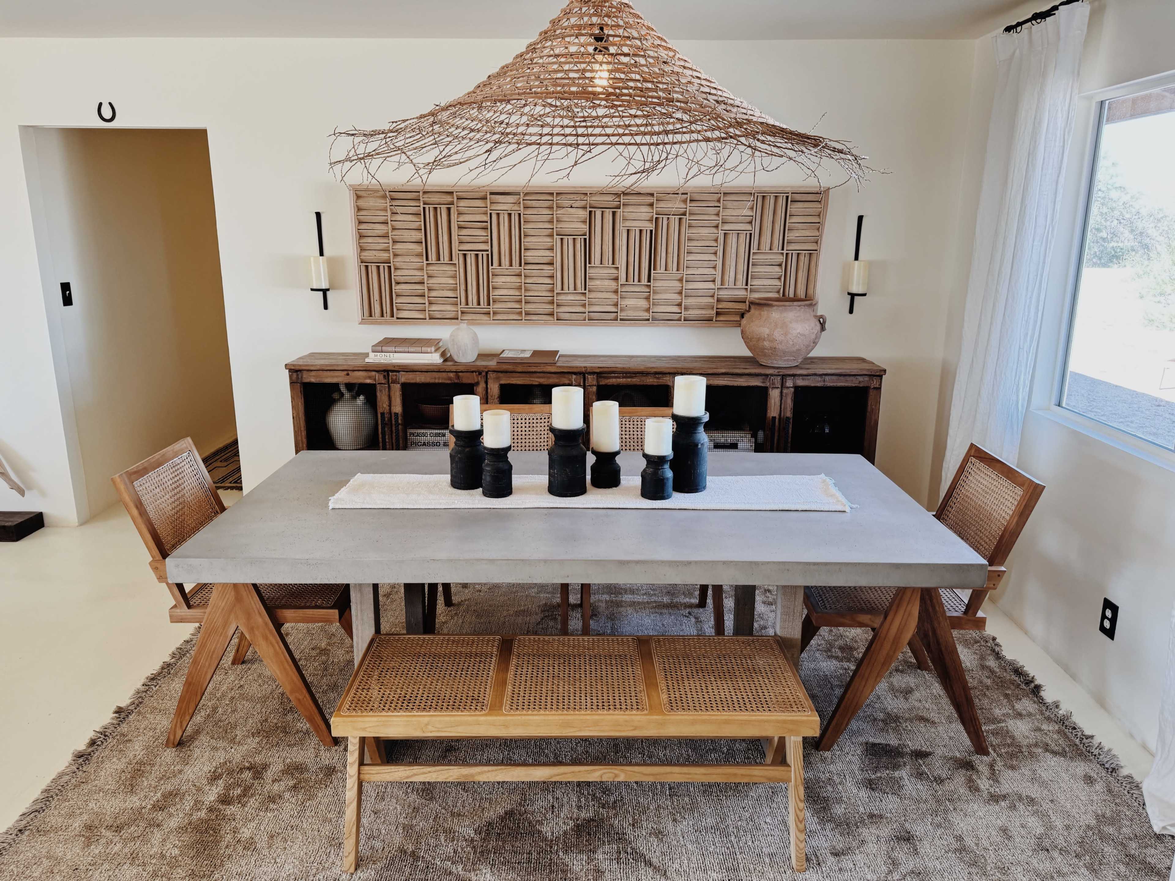 The image features a modern dining area with a concrete table, wooden chairs, a woven pendant light, and a decorative bamboo wall behind a sideboard.