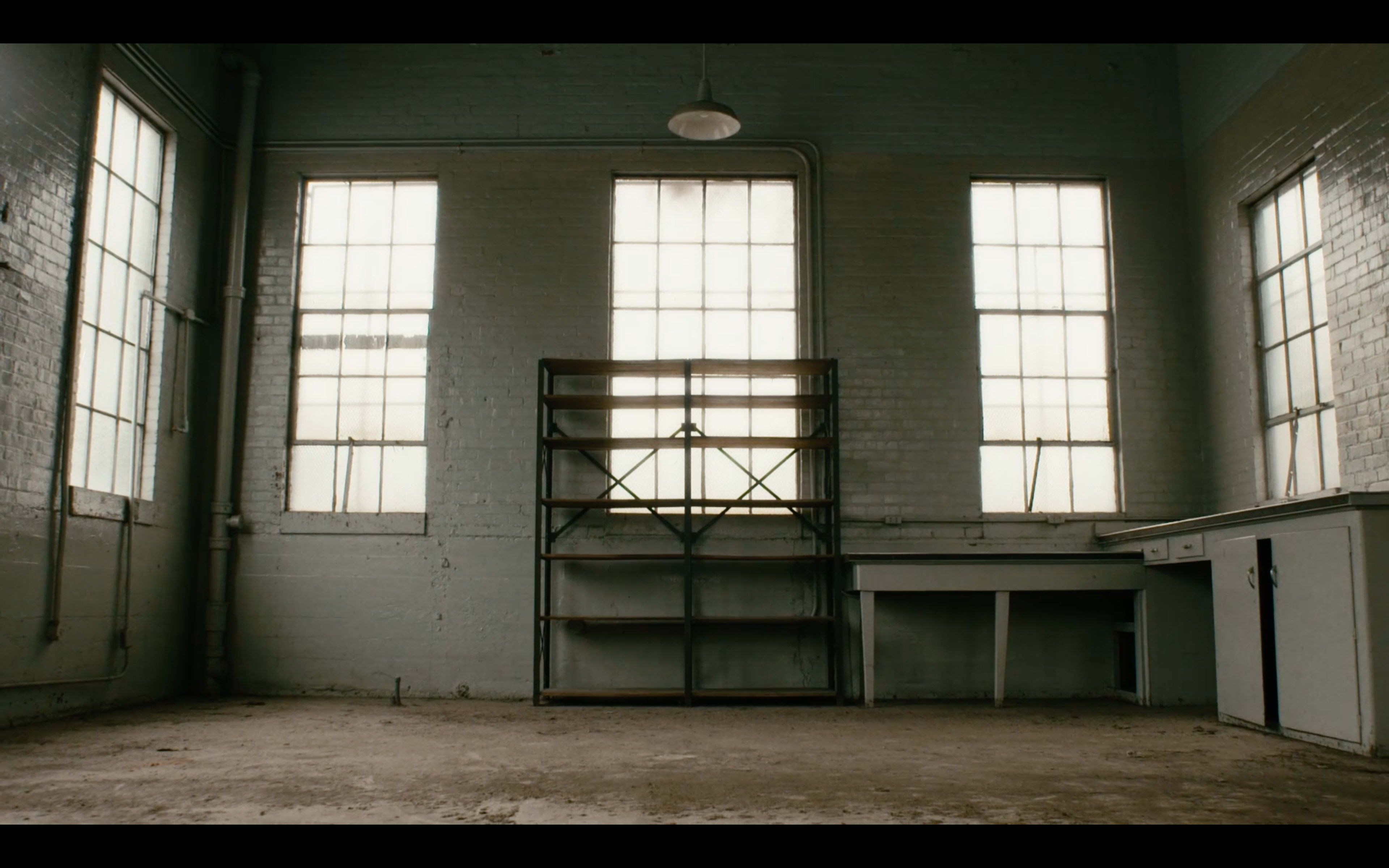 An empty, well-lit room with large windows, a metal shelving unit against one wall, and a small table with drawers.