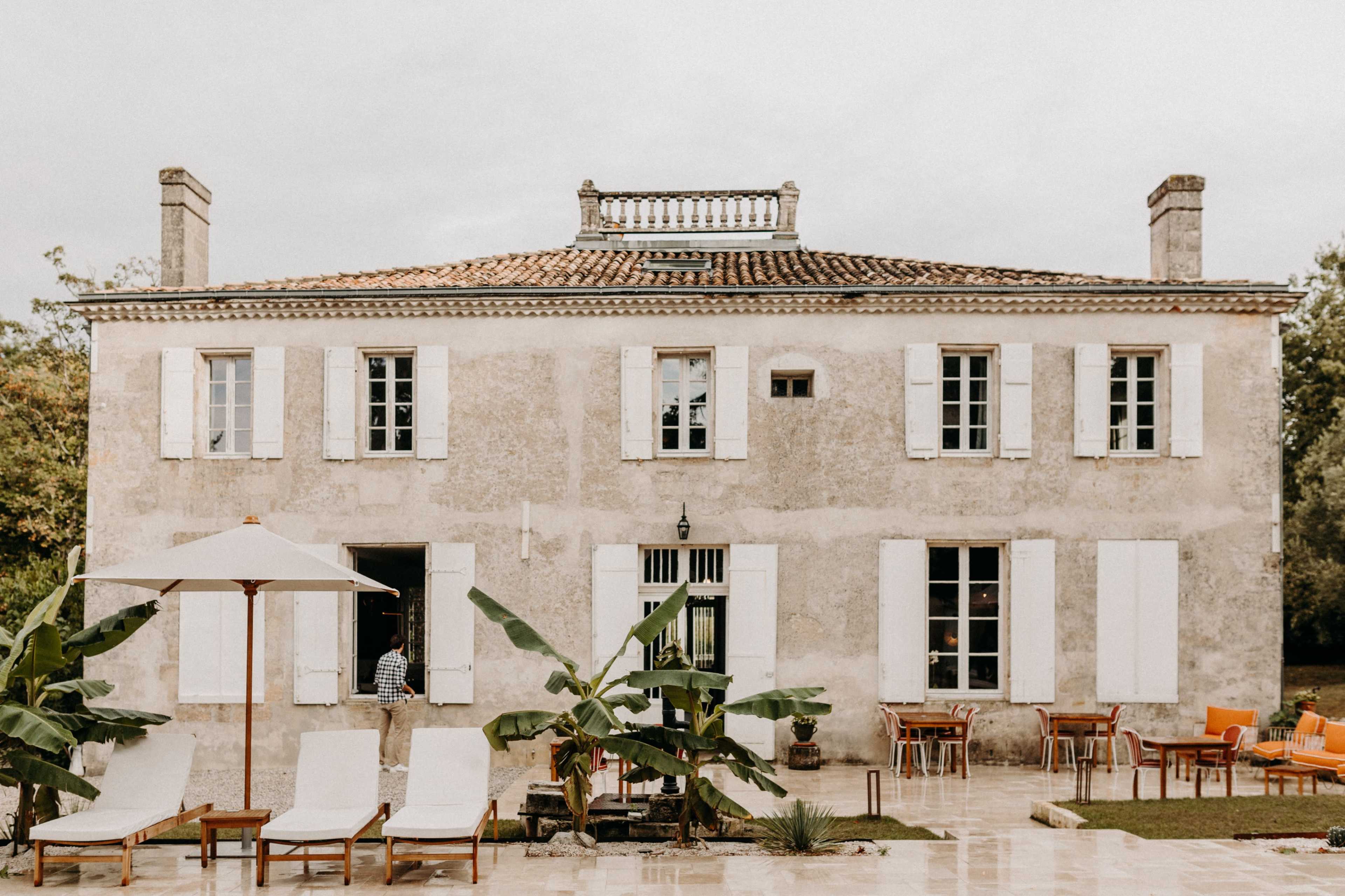 A weathered two-story house with white shutters, a terrace with lounge chairs, and an umbrella, surrounded by greenery.