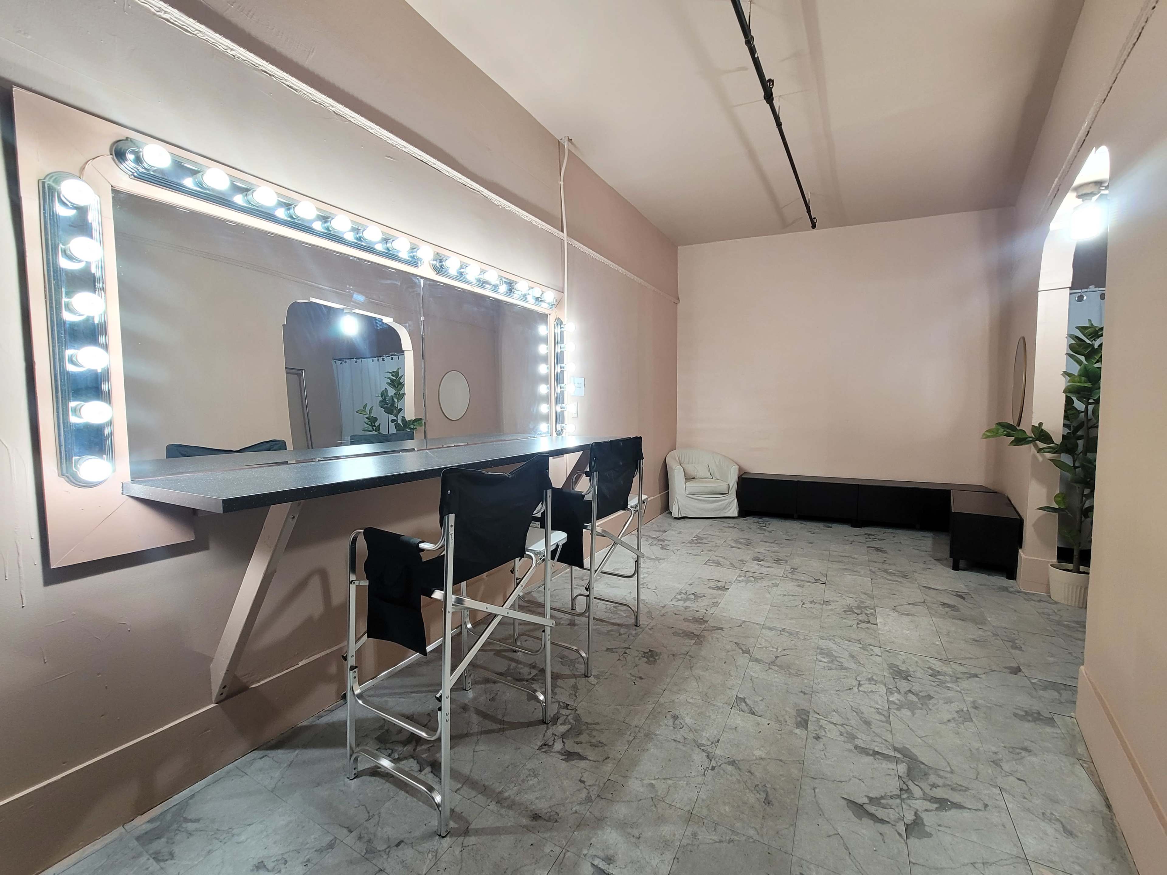 The image shows a minimalistic dressing area featuring a long mirror illuminated by bright lights, a row of black stools, and a white armchair against a light-colored wall with a marble floor.