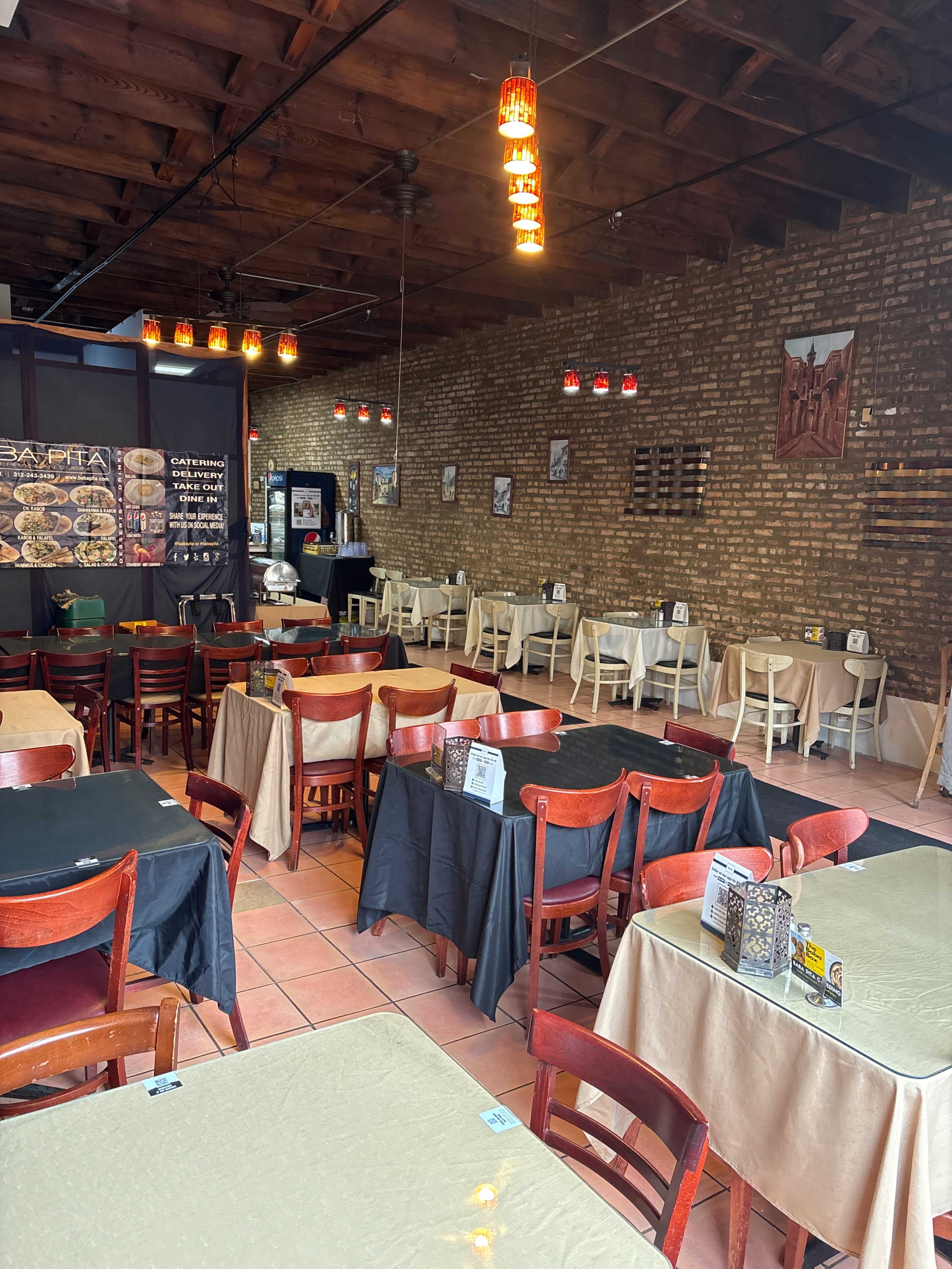 The image shows an empty restaurant featuring wooden tables with black and beige tablecloths, exposed brick walls, and hanging light fixtures.