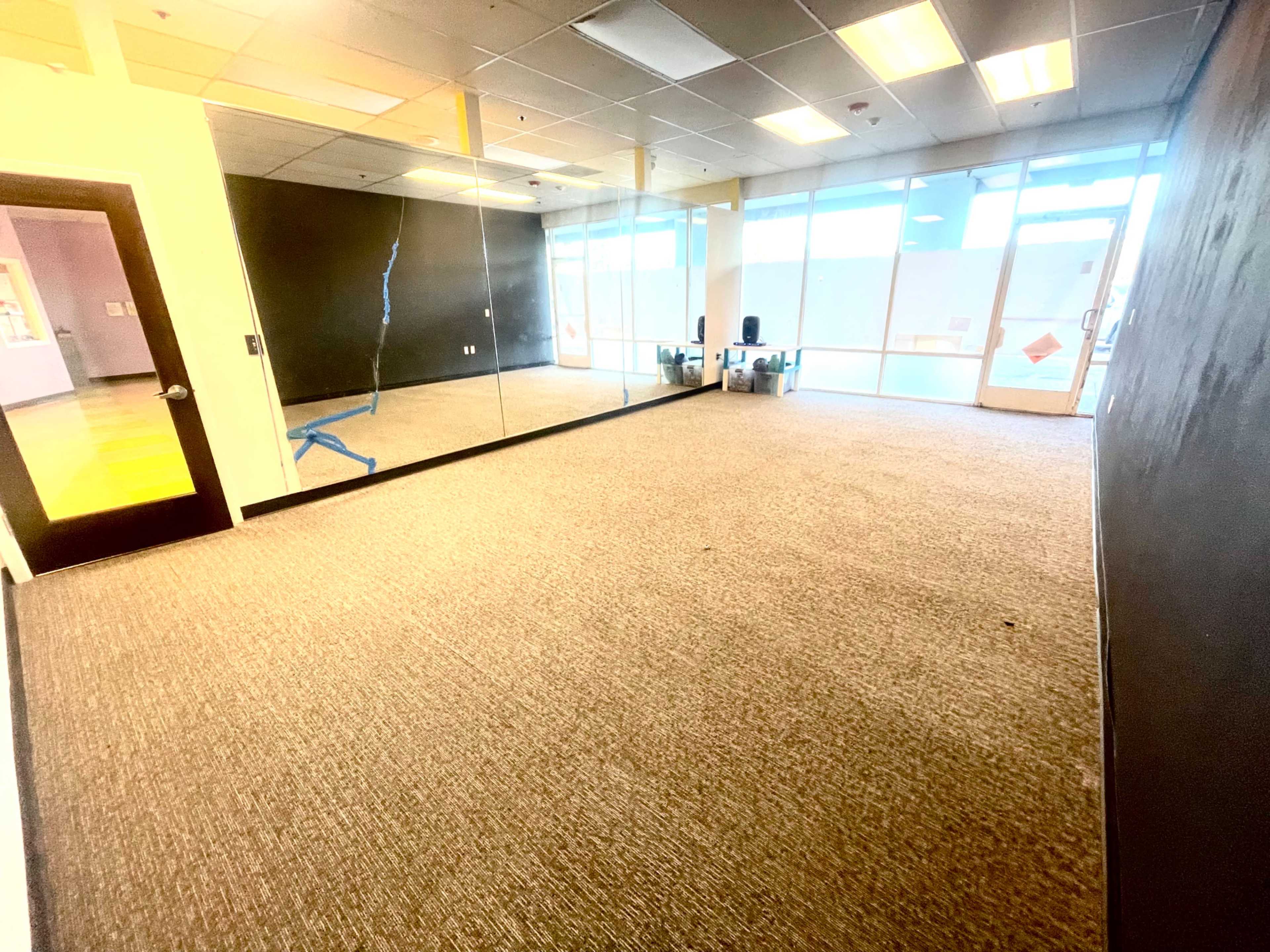 A spacious room with carpeted flooring, large windows allowing natural light, and a mirrored wall on one side.
