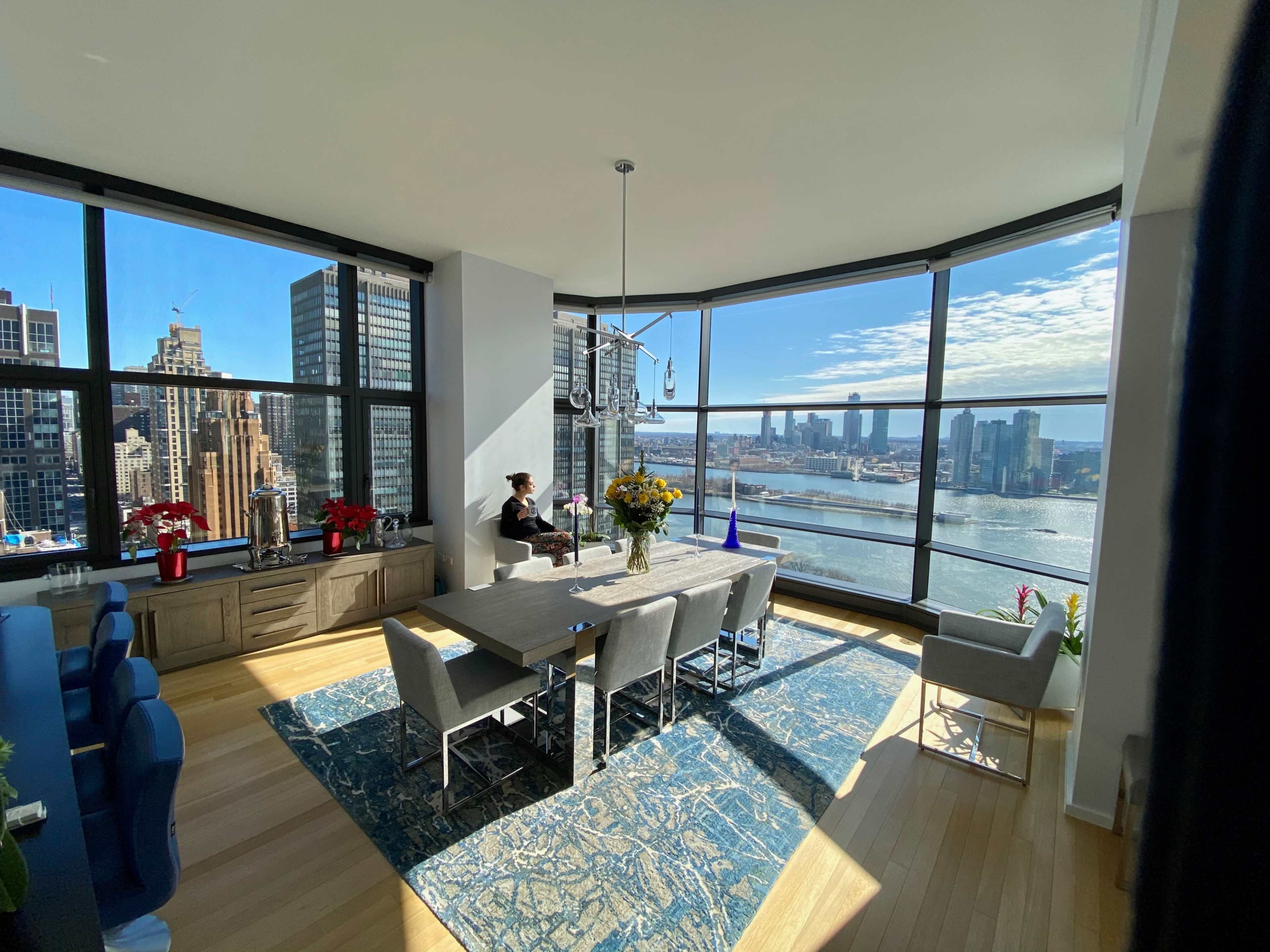 A dining area with a large table and chairs overlooks a cityscape through expansive windows, showcasing a river and skyline.