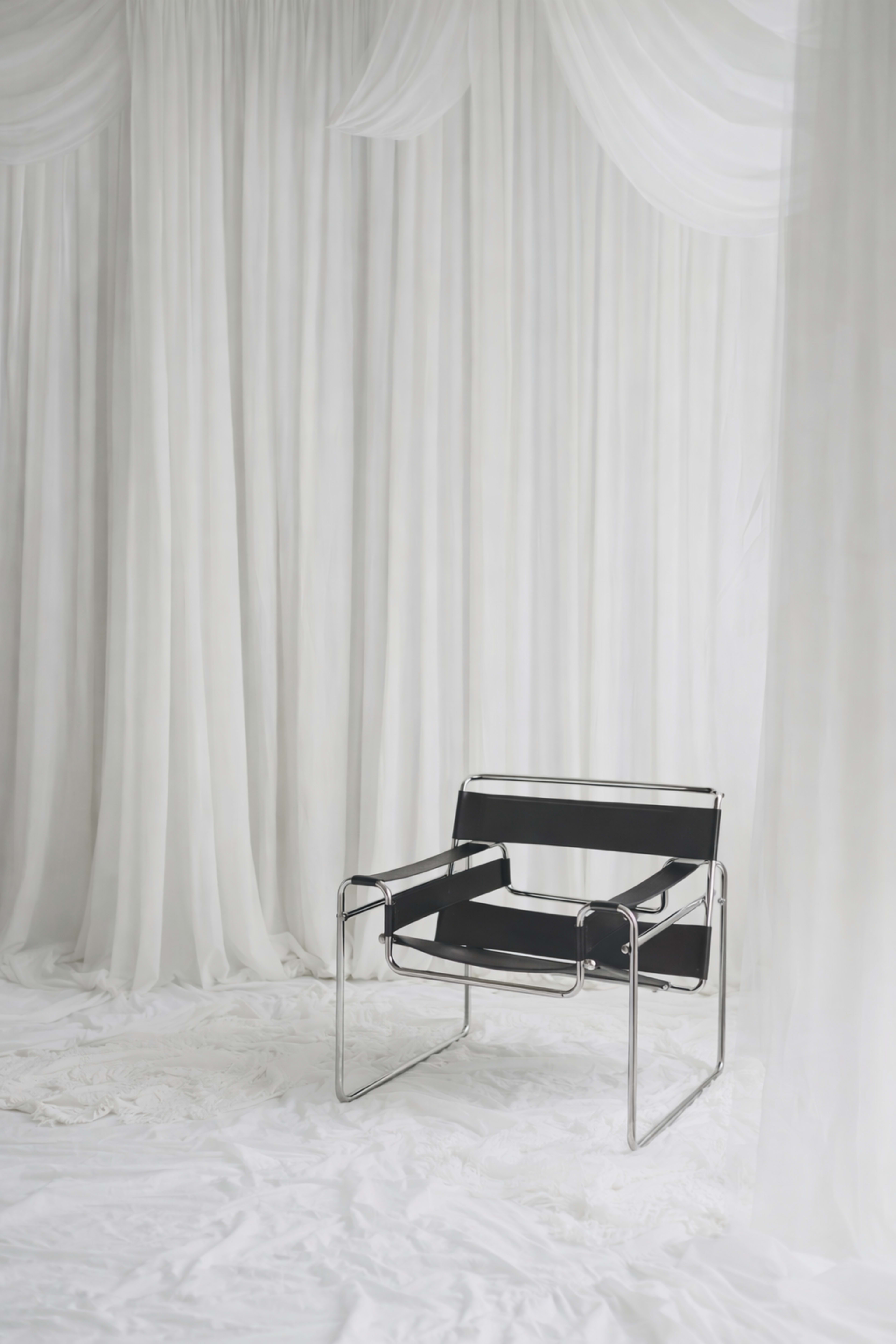 A black chair with a metal frame is positioned in front of sheer white drapes.