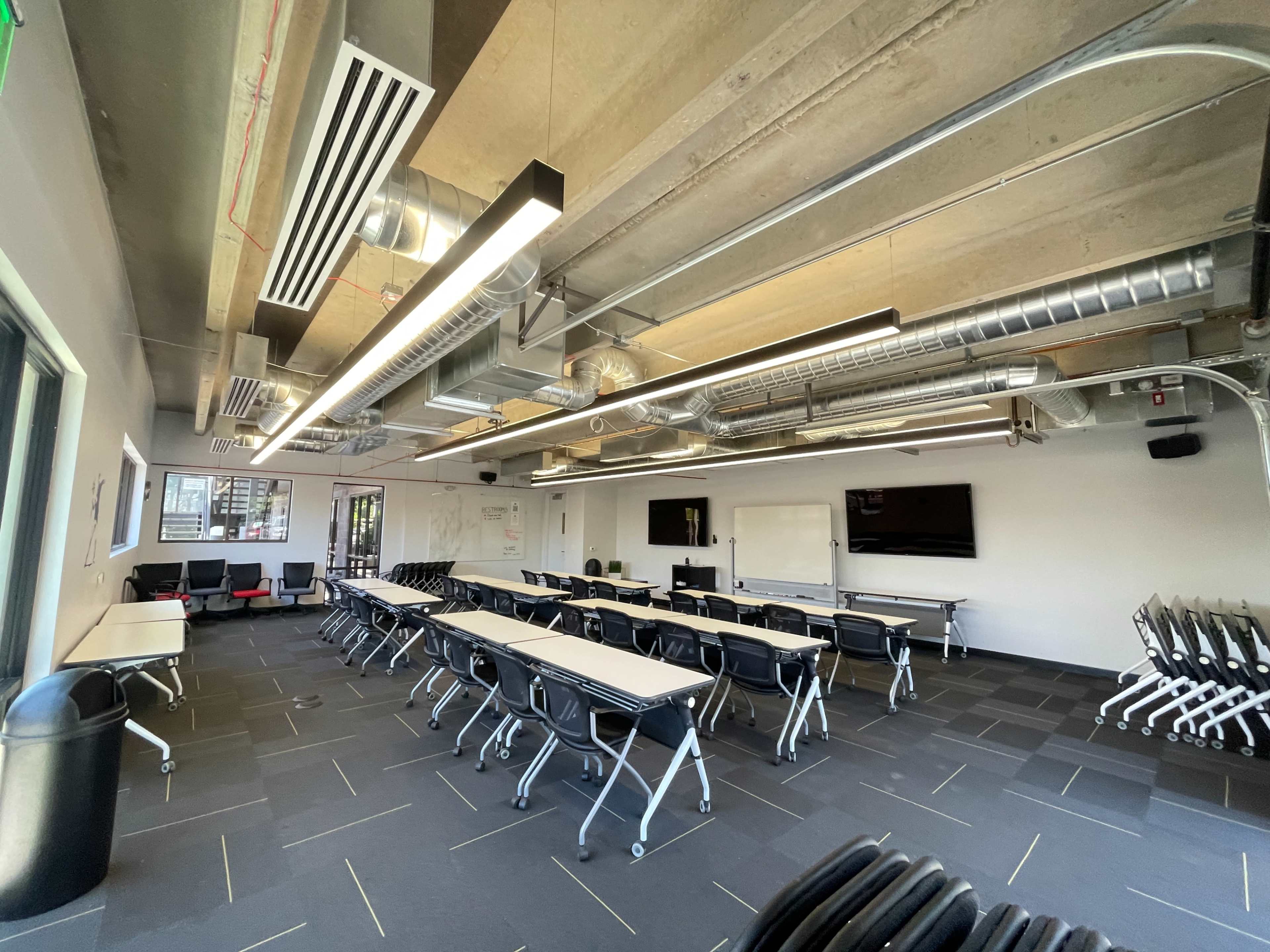 A modern conference room with rows of tables and chairs arranged neatly, large windows, and exposed ductwork on the ceiling.