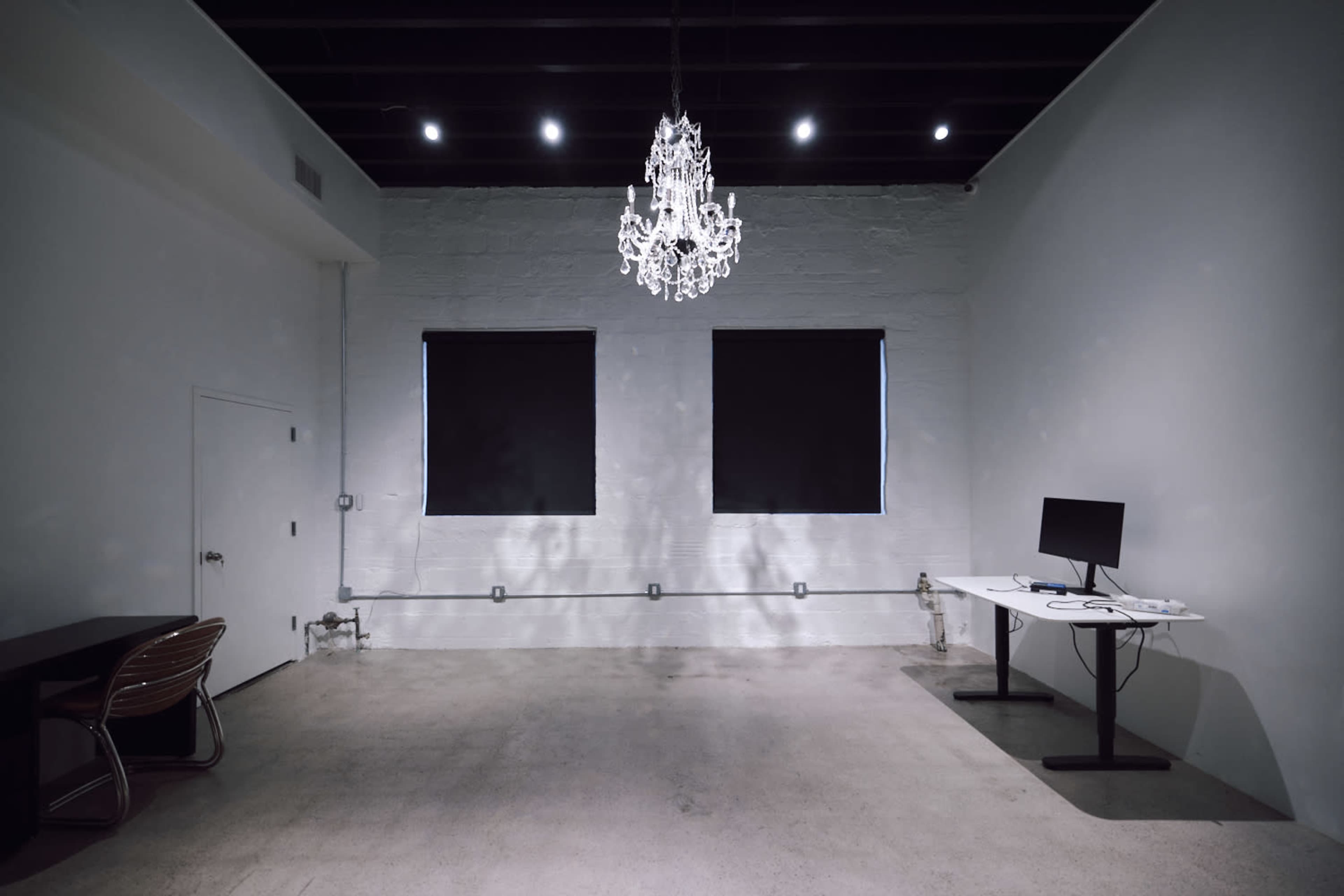 The image shows a minimalistic room with a chandelier, two windows with dark blinds, a desk, and a monitor on a standing desk.