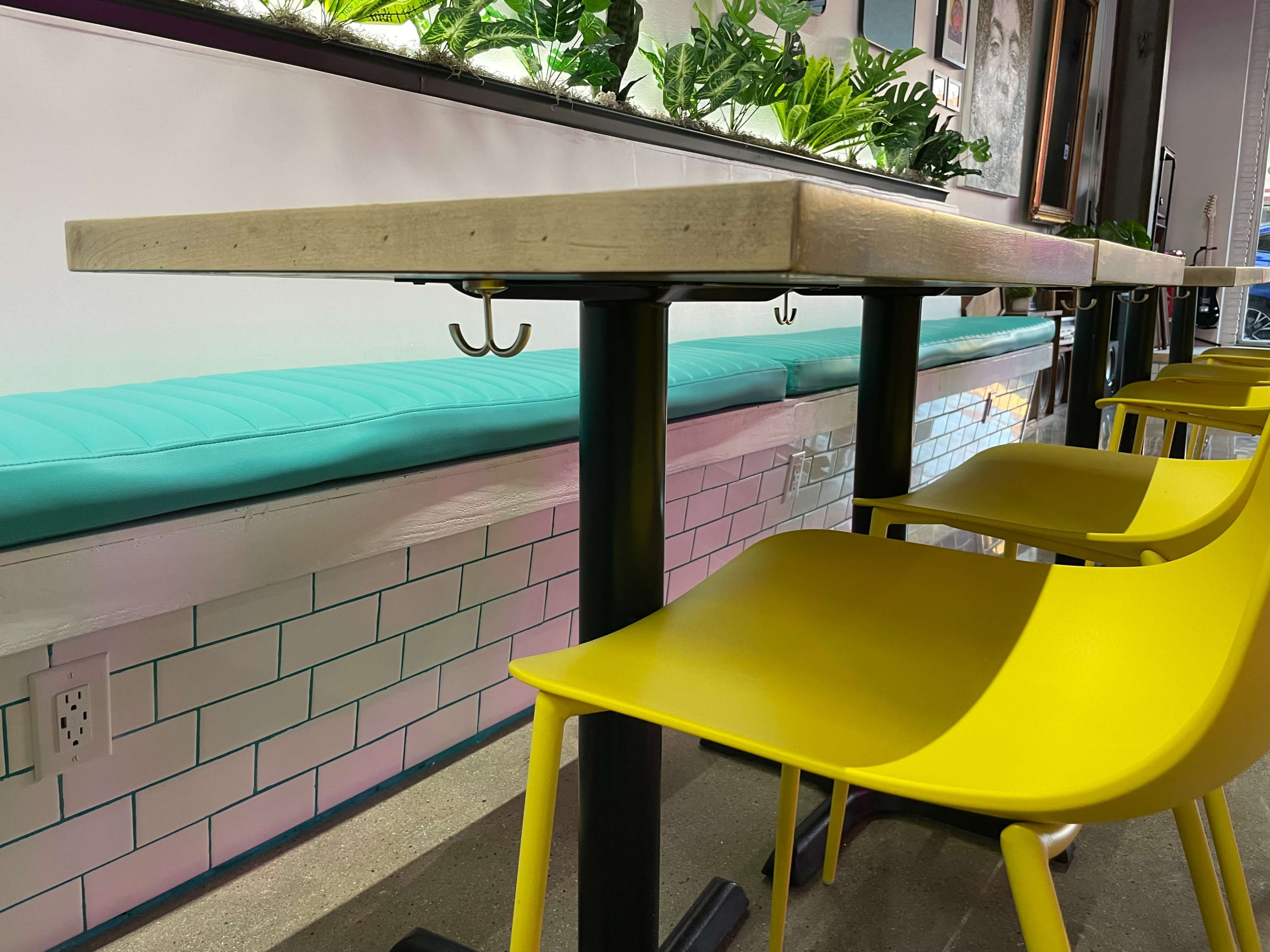 A light wooden table with a metal base and yellow chairs, positioned against a wall with a turquoise bench and decorative plants.