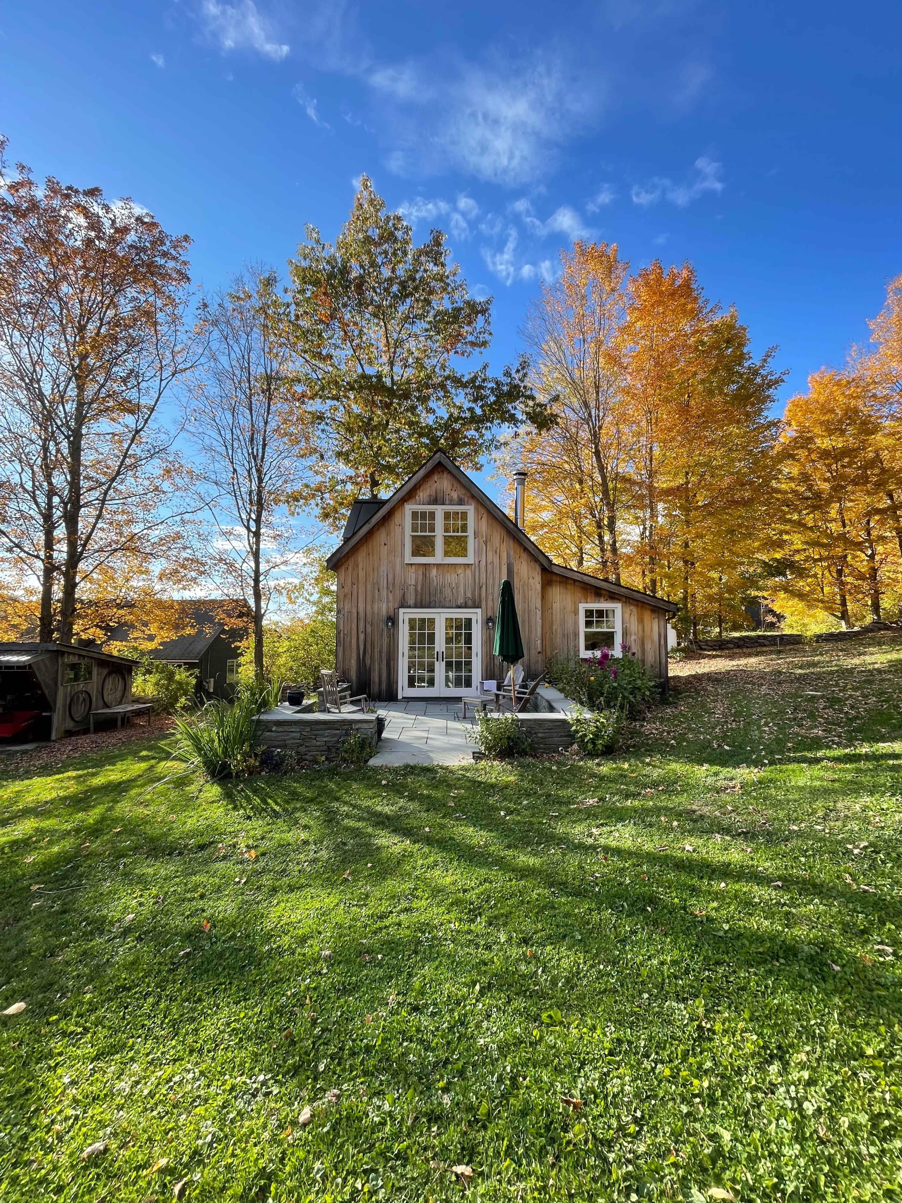 A wooden house with a sloped roof stands amid colorful autumn trees, surrounded by a grassy area and a stone patio.
