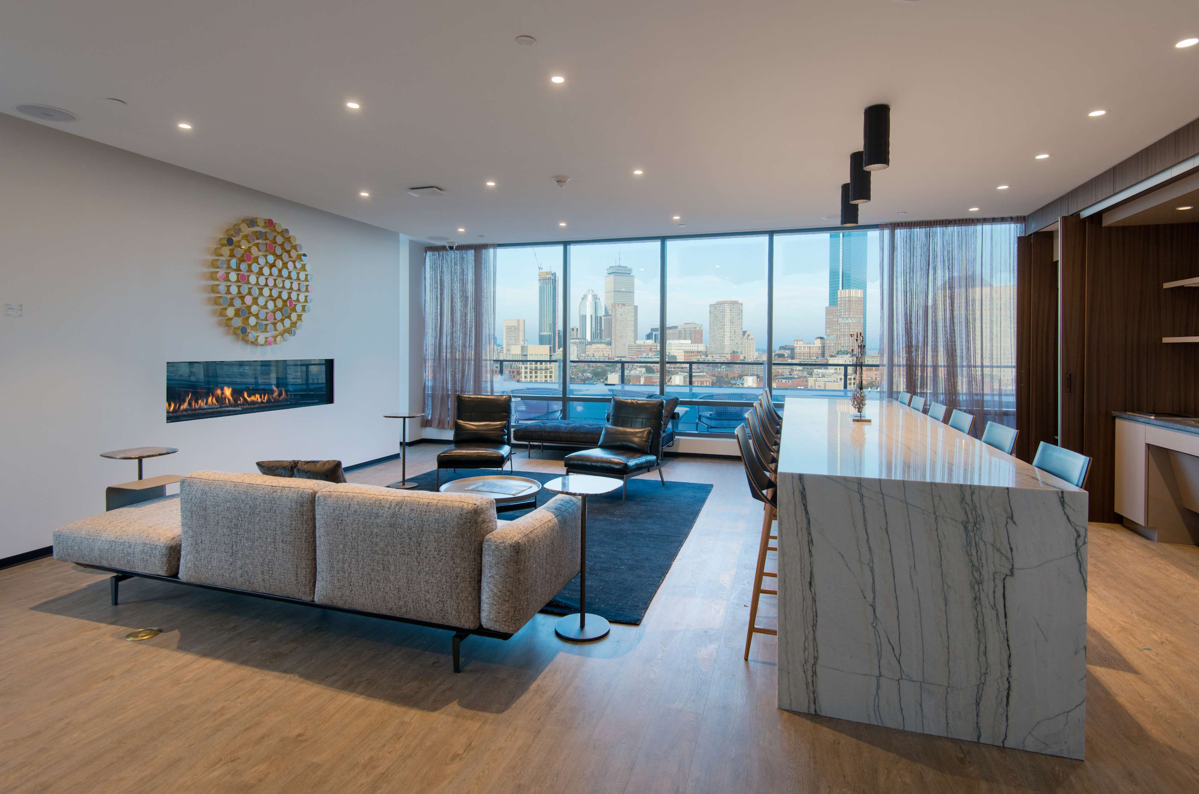 A modern living room features a large window with city views, accompanied by a sleek sofa, chairs, a marble-topped bar, and a decorative wall piece.
