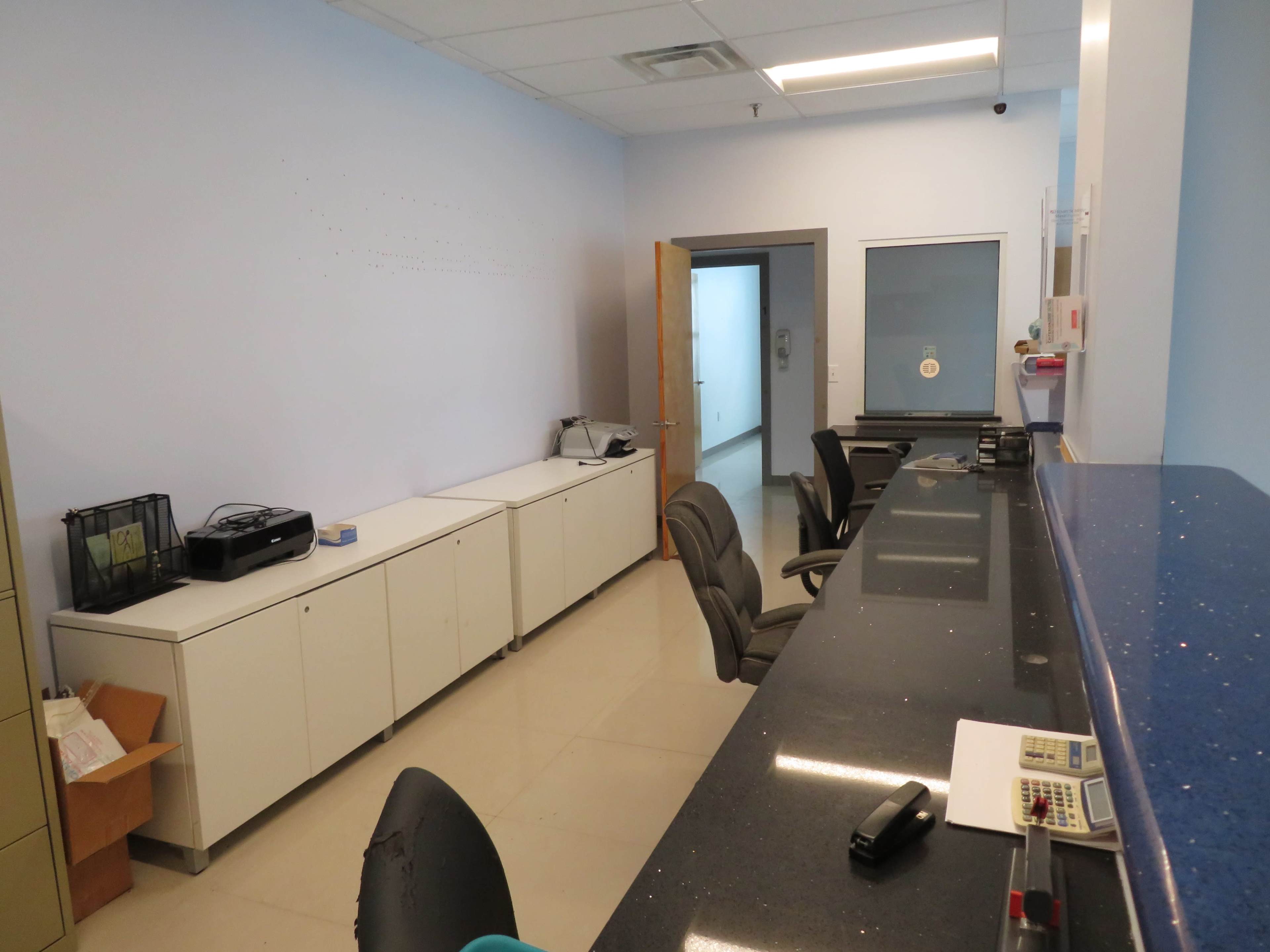 The image shows a reception area with a long counter, several office chairs, and filing cabinets against the walls in a room with light blue walls and a door leading to another space.