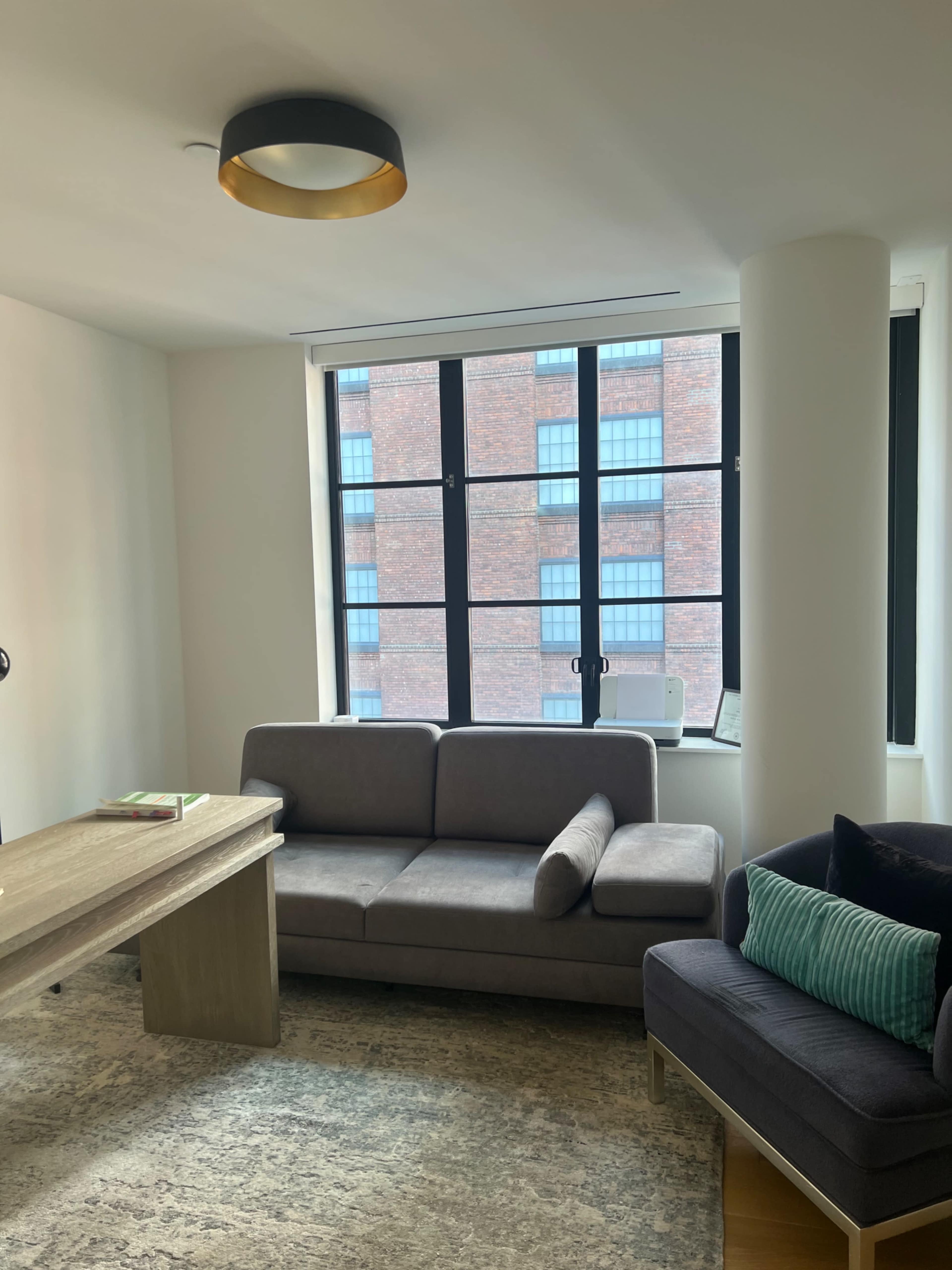 The image shows a modern living room with a gray couch, a dark armchair, a wooden coffee table, and large windows revealing a brick building outside.