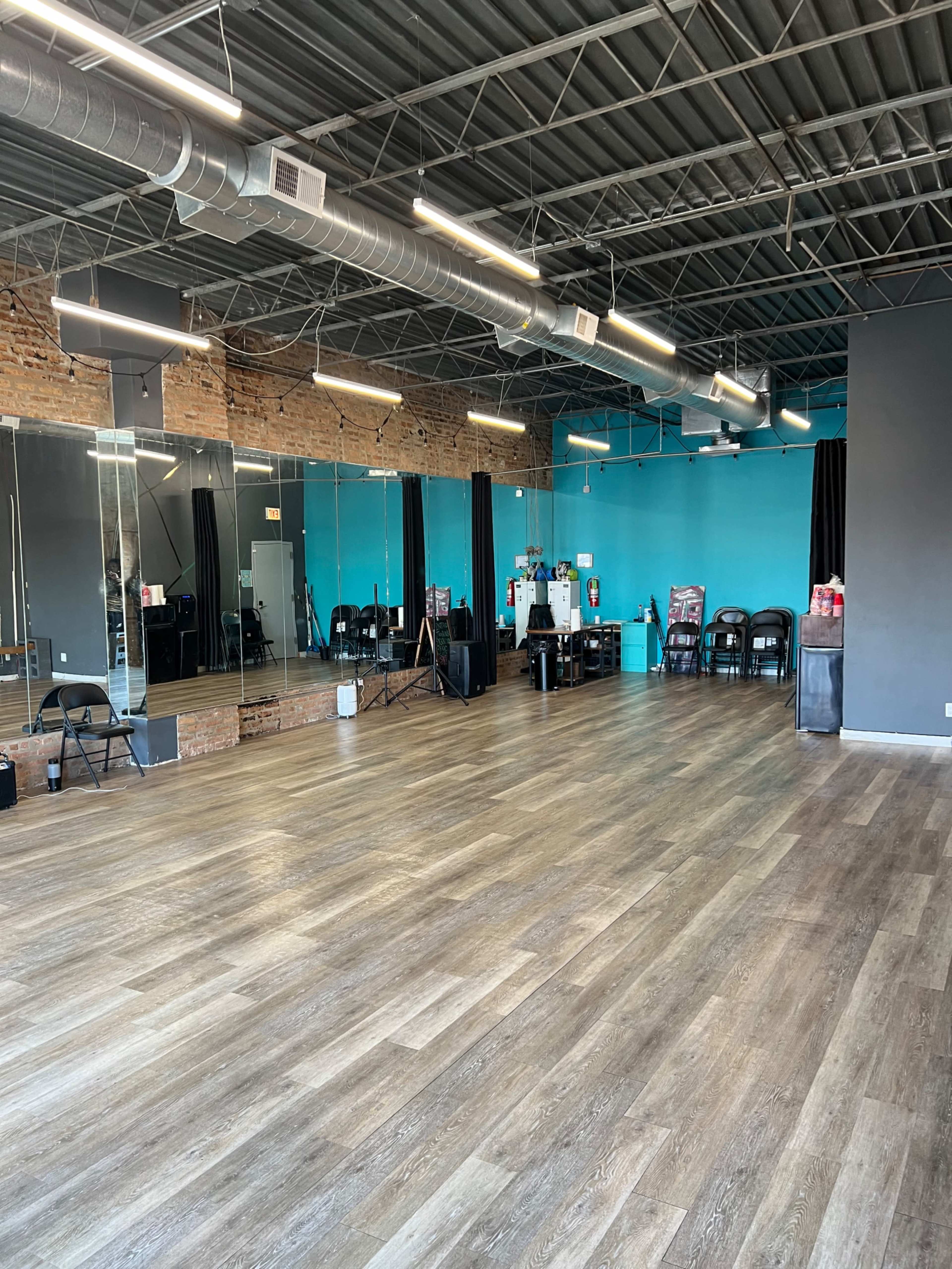 The image shows a spacious dance studio with wooden flooring, mirrored walls, and a bright blue accent wall.