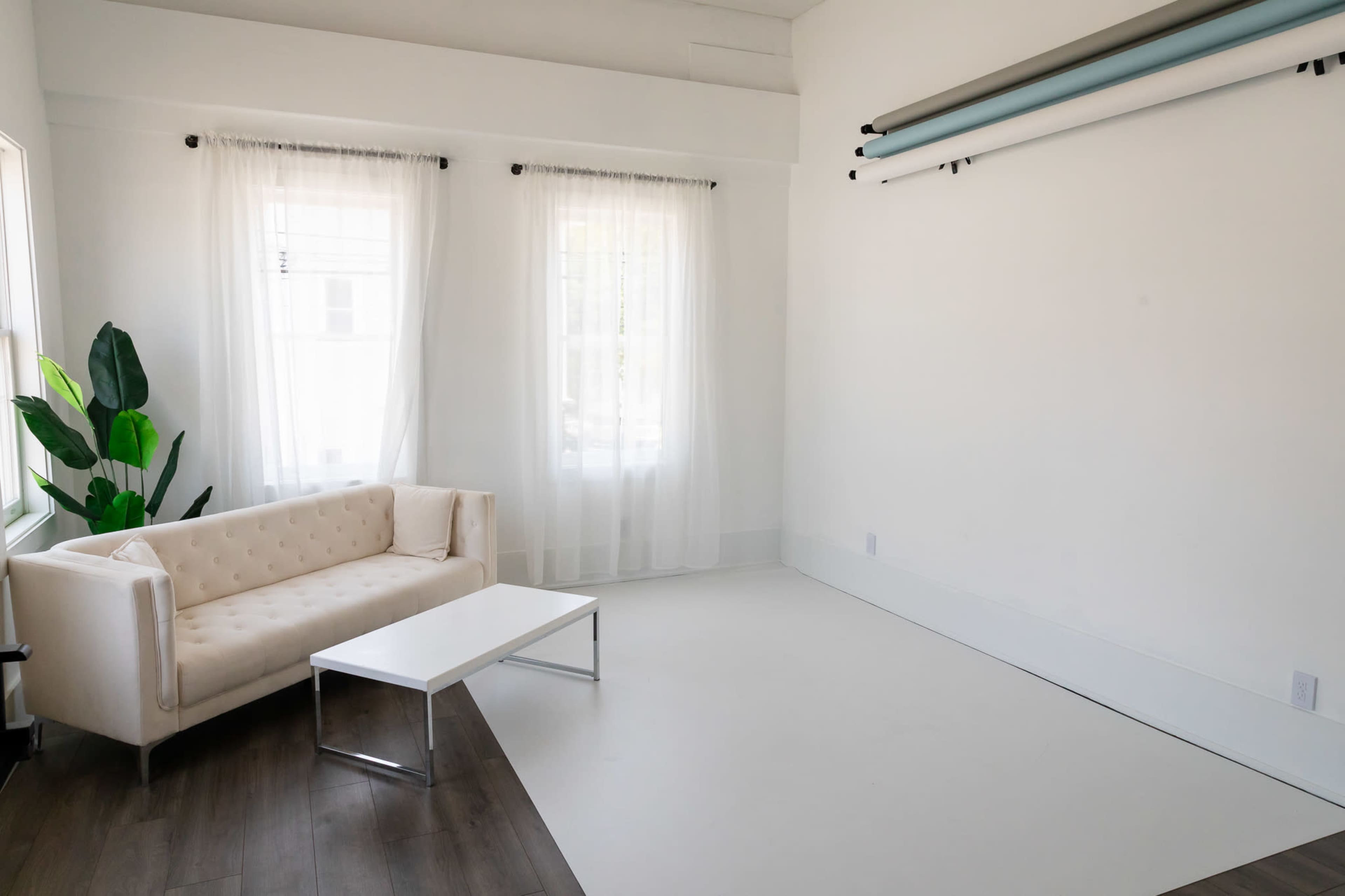 The image shows a bright, minimalistic studio space featuring a white couch, a rectangular coffee table, and large windows with sheer curtains.