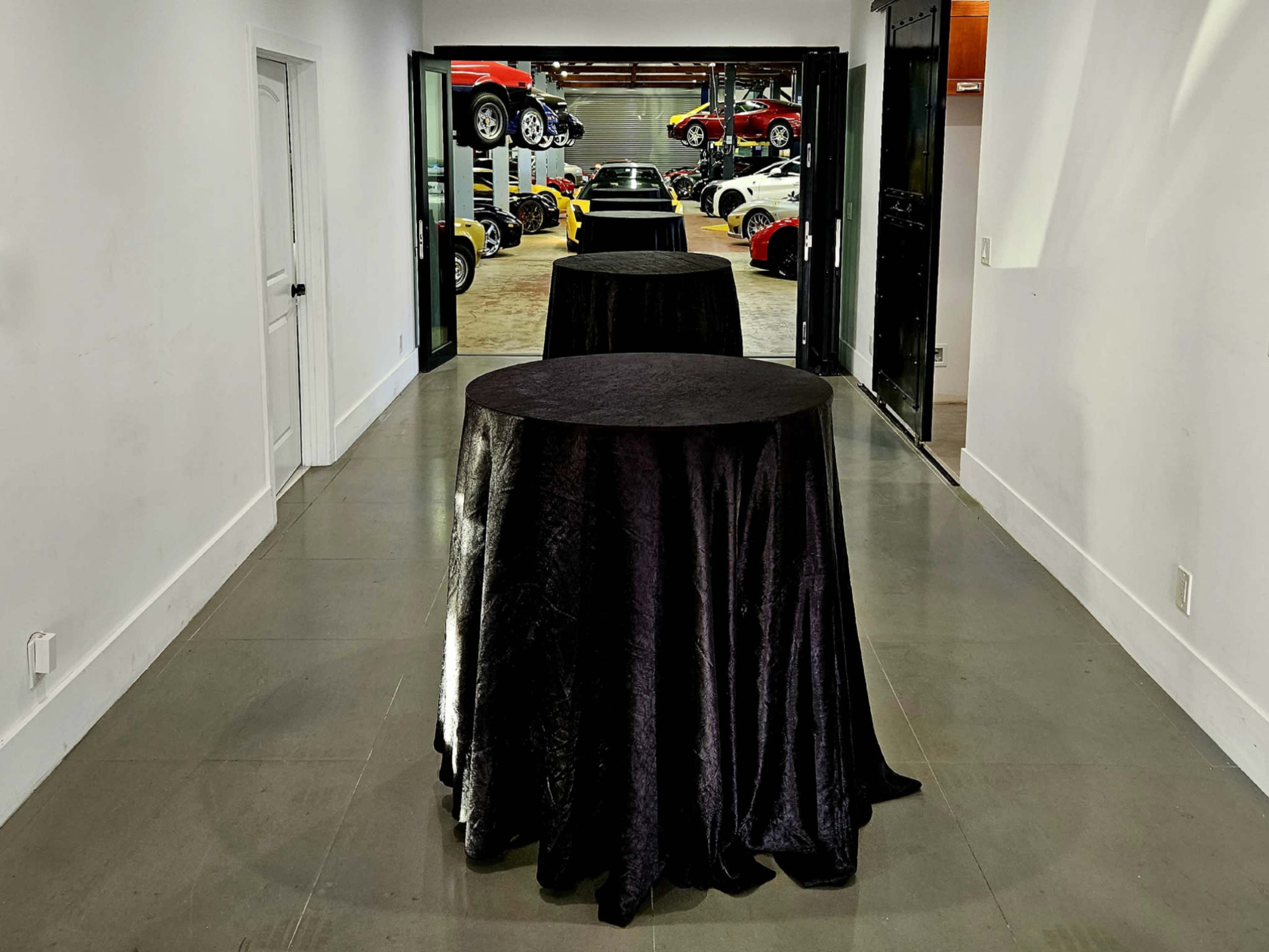 Three black-draped tables are positioned in a narrow hallway leading to a garage filled with various cars.