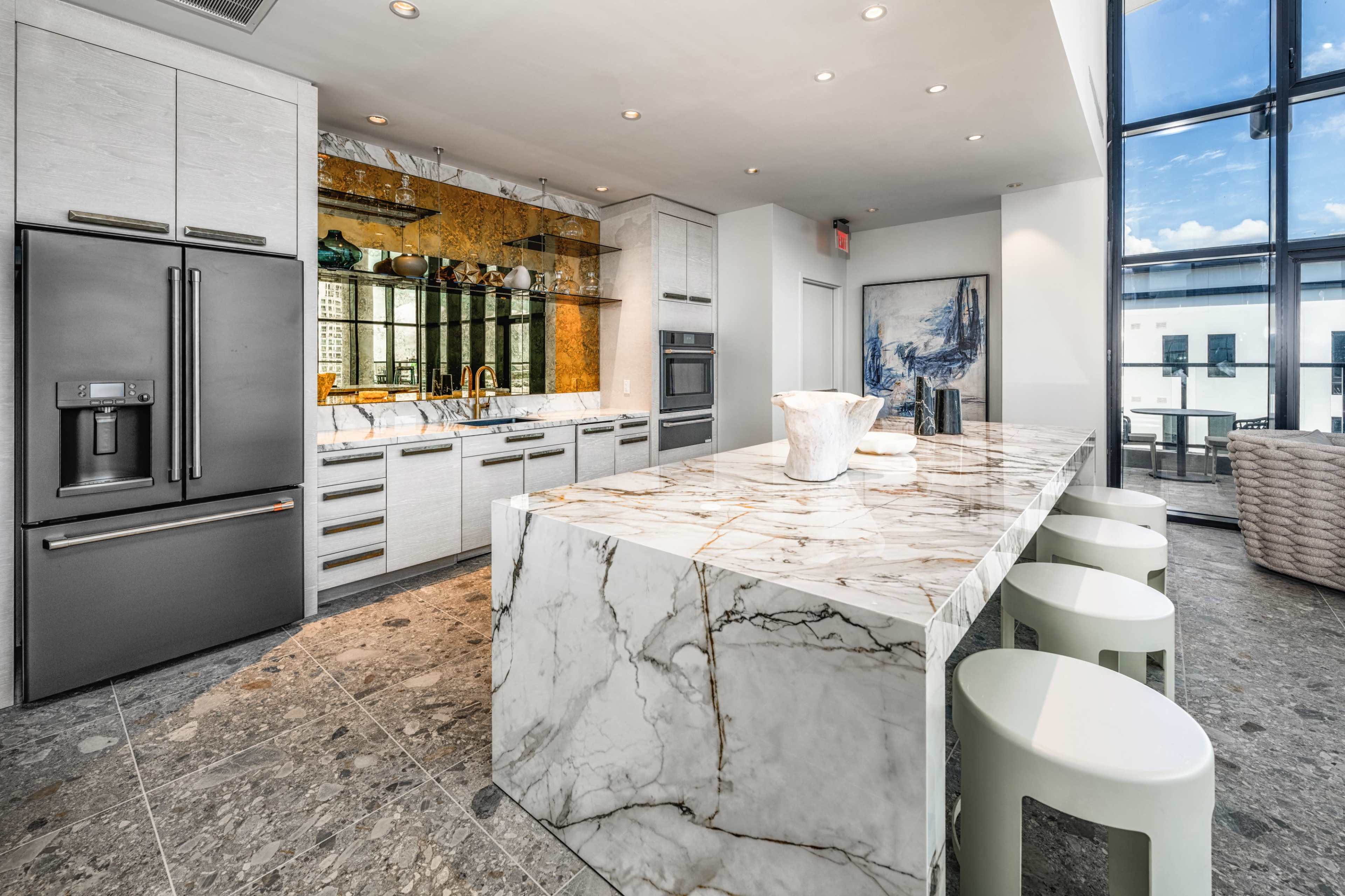 The image shows a modern kitchen featuring a large marble countertop, stainless steel appliances, and ample natural light from large windows.