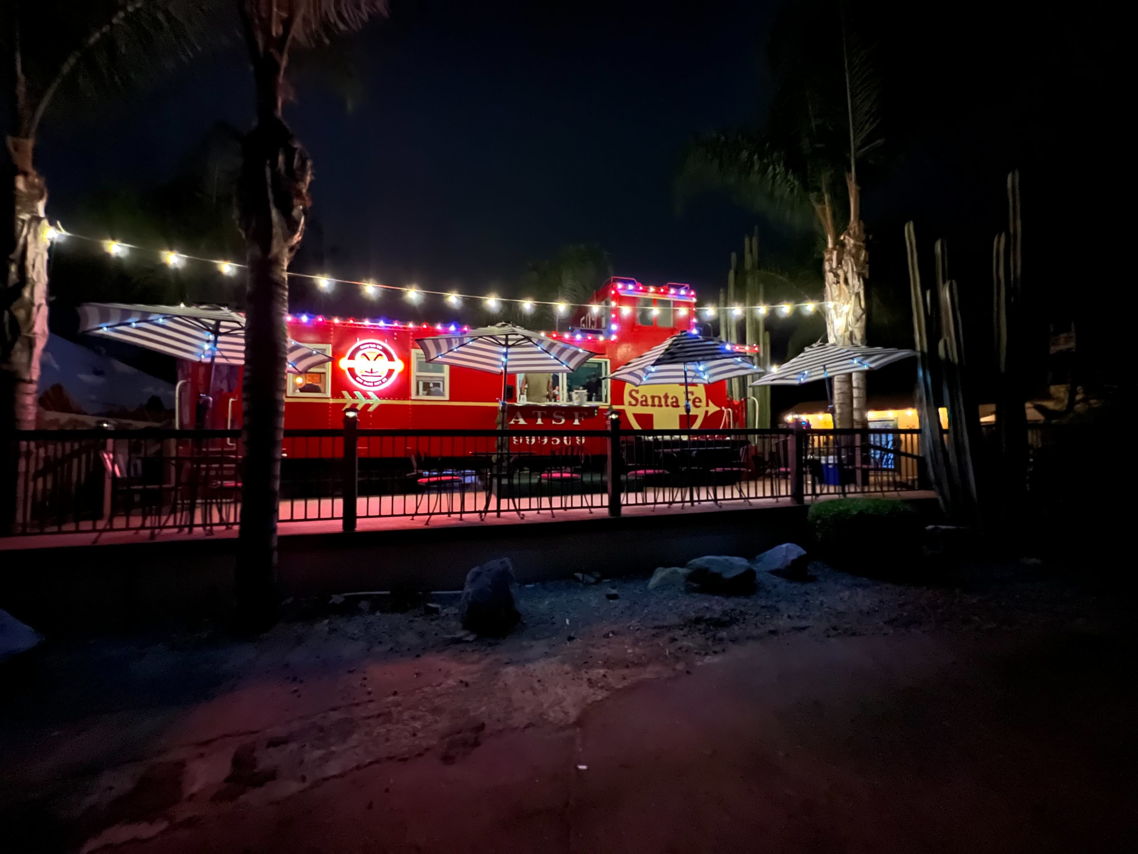 A brightly lit food truck with colorful lights and striped umbrellas is set up in a garden-like area at night.