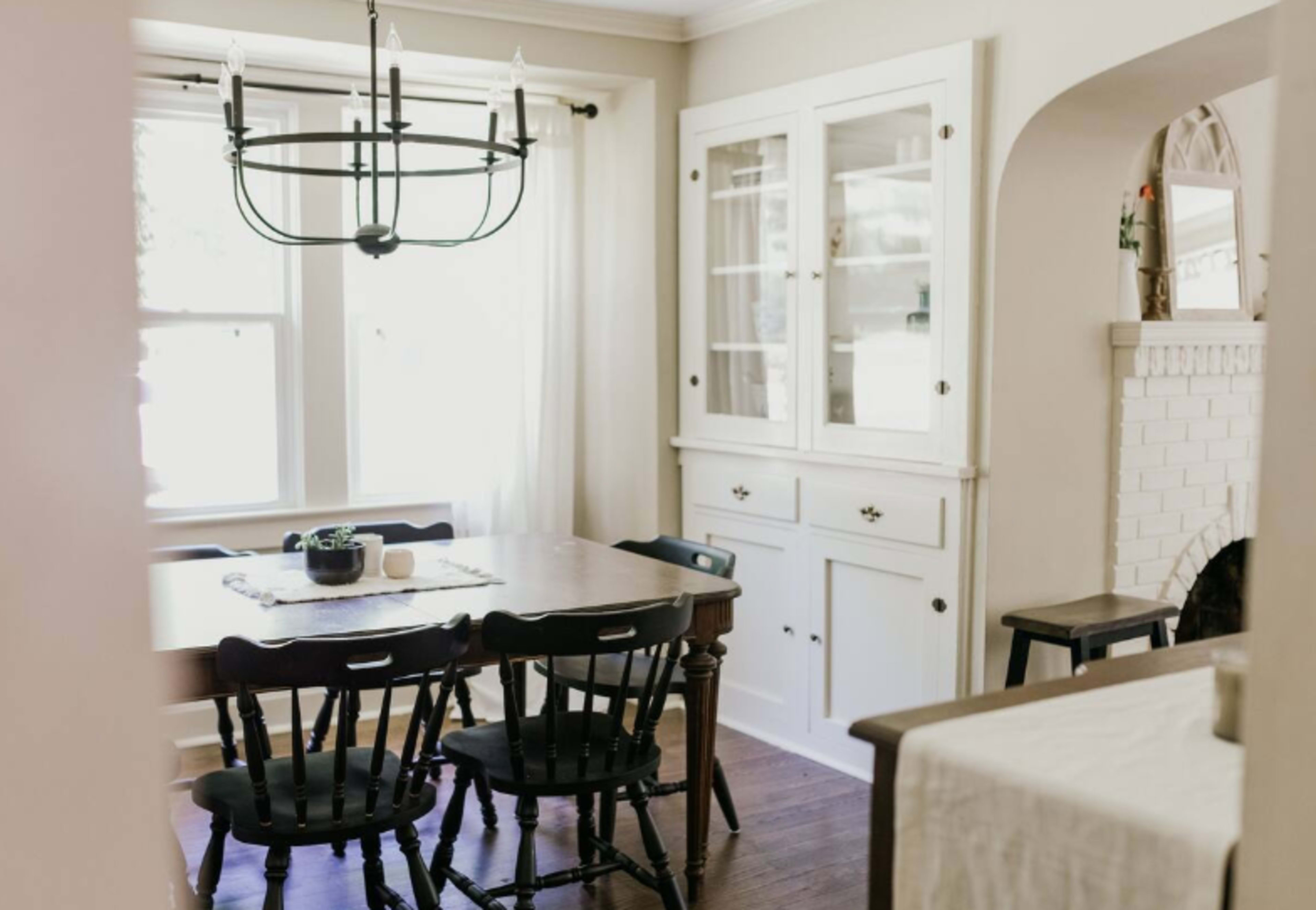 A dining area features a table surrounded by black chairs, a chandelier above, and a white cabinet against the wall.
