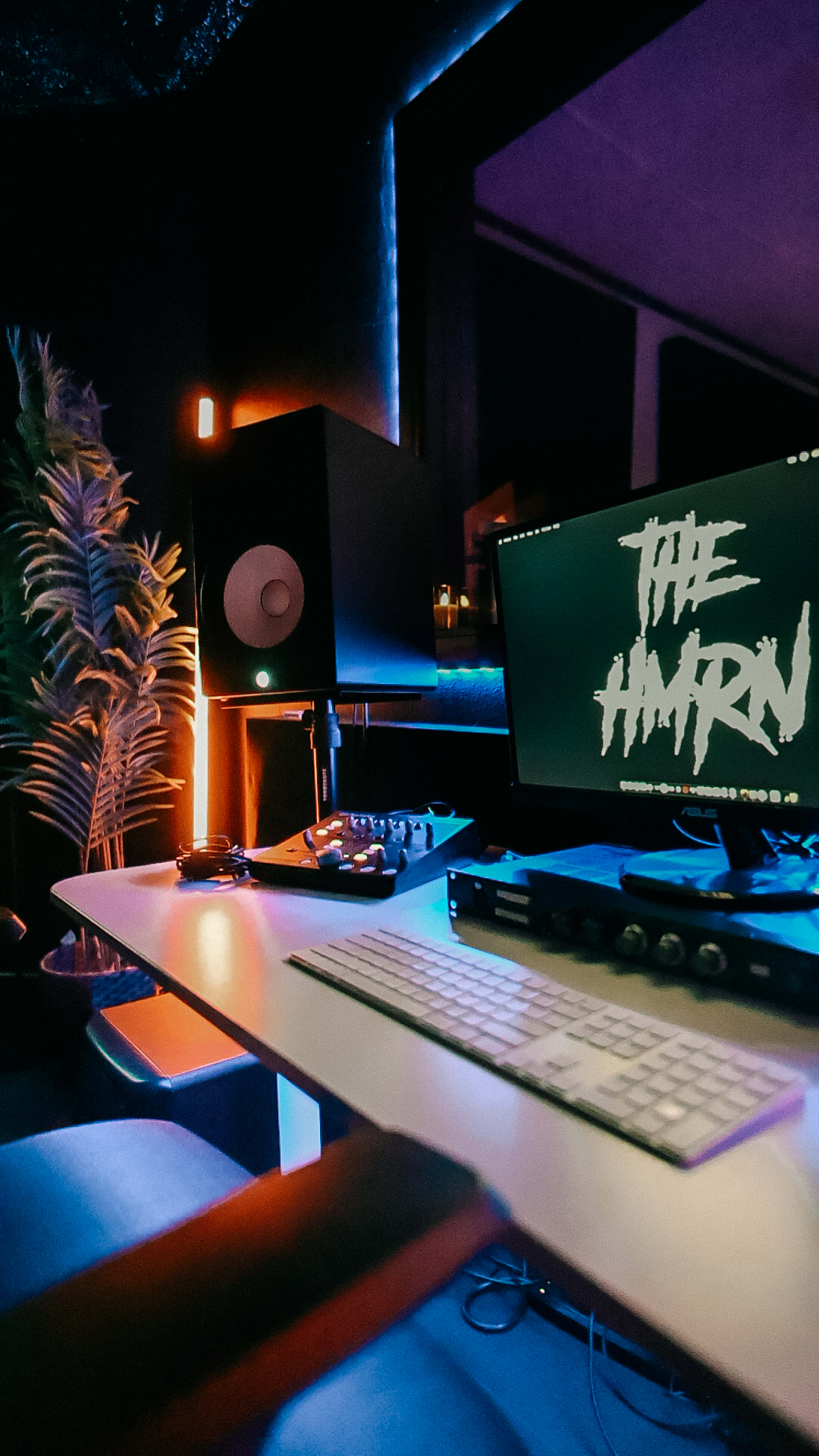 A modern music production studio features a sleek desk with a computer screen displaying "THE AMRN," studio monitors, a keyboard, and DJ equipment, all illuminated by ambient lighting.