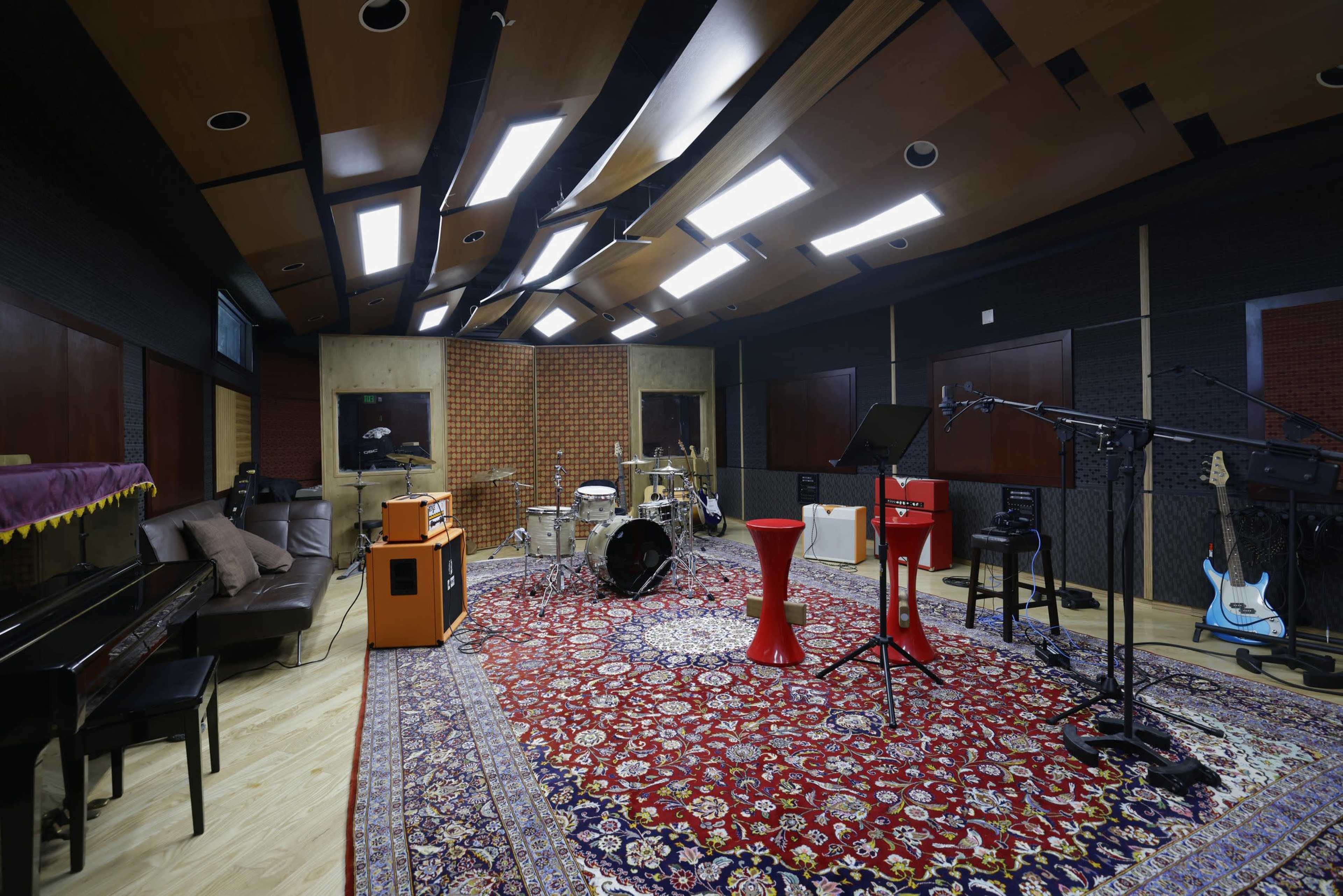 The scene shows a music recording studio featuring a drum set, various instruments, and red accent tables on a large area rug.