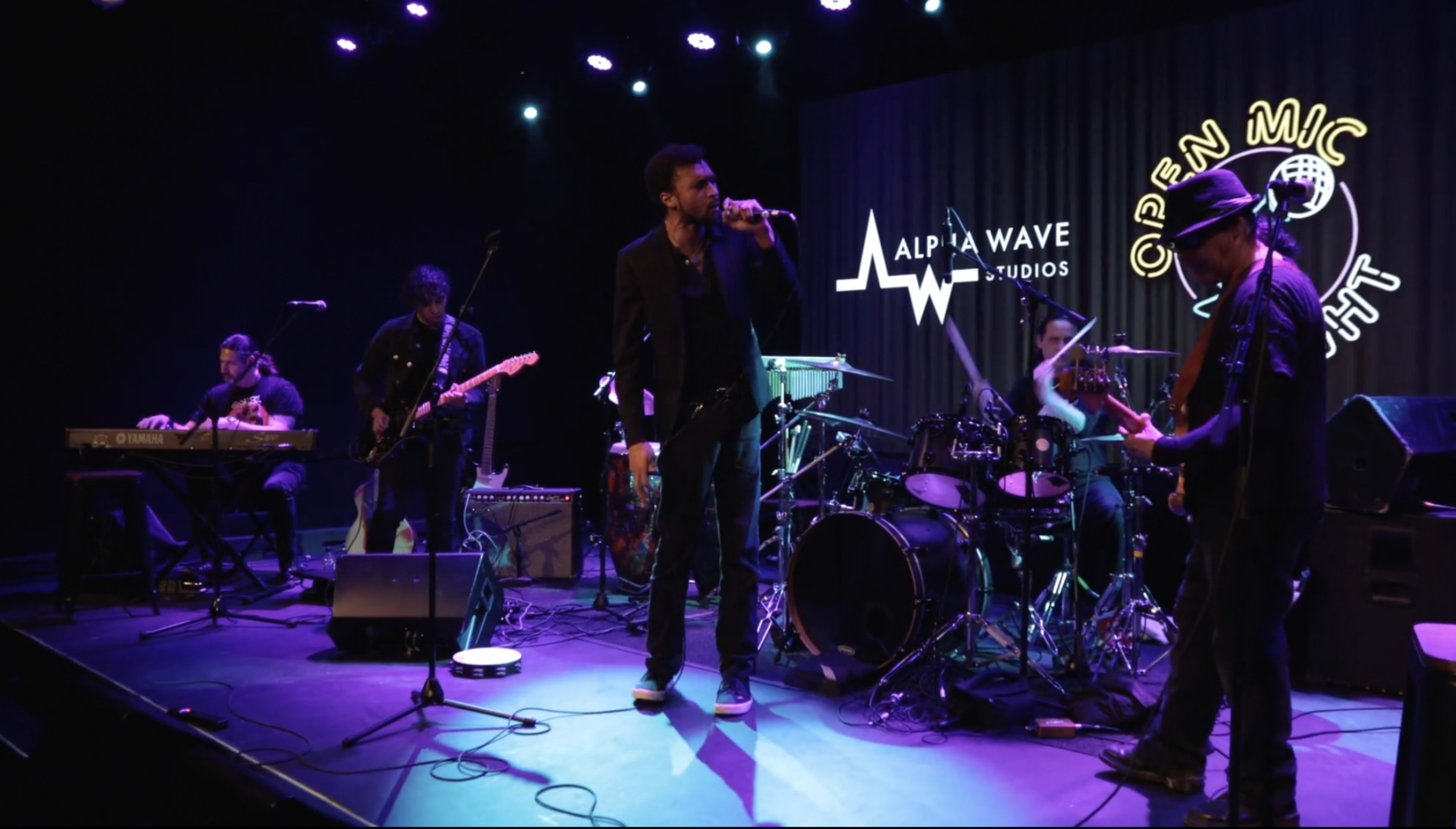 A band performs on stage at Alpha Wave Studios during an open mic night, with various instruments and musicians engaged in the performance.