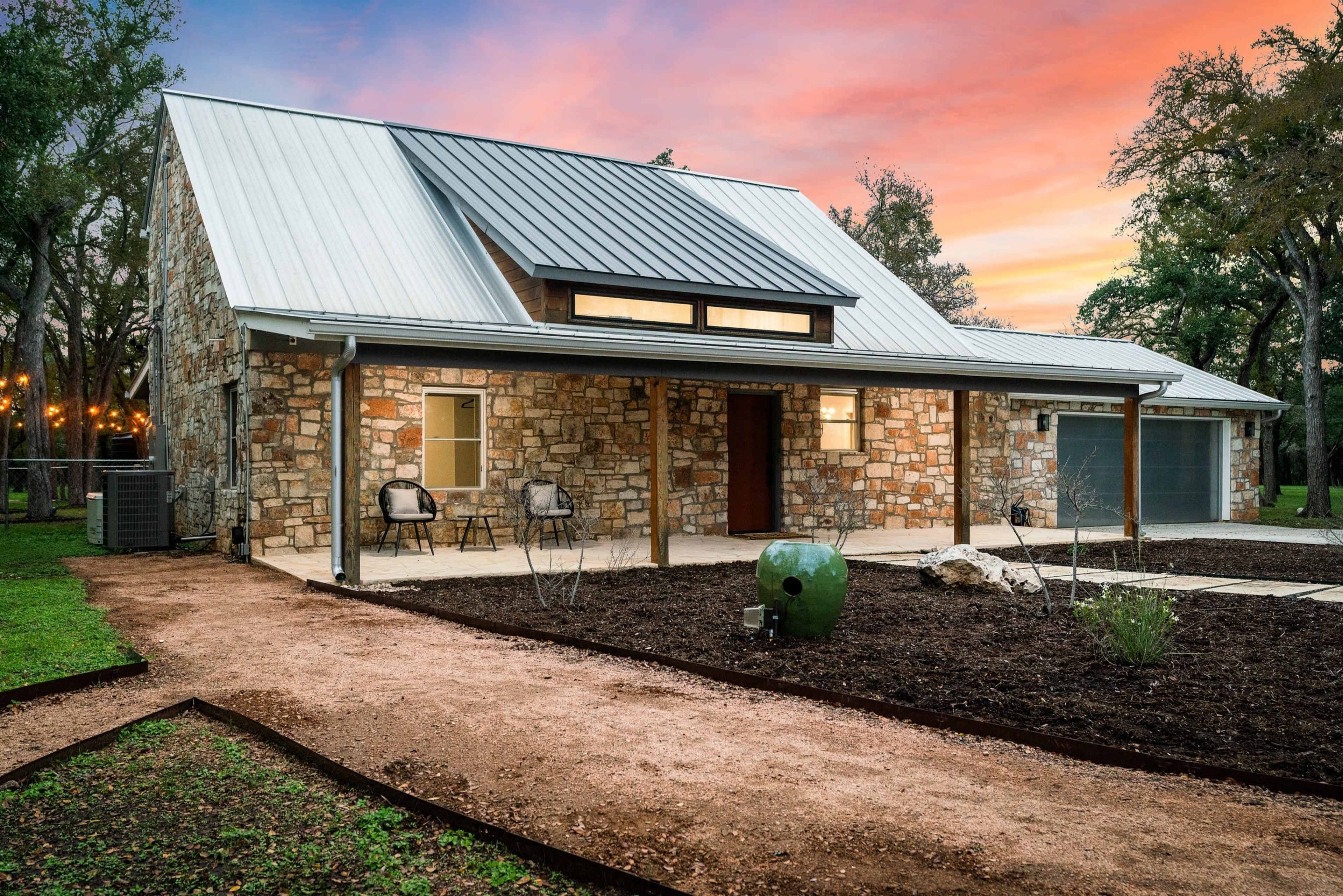 The image shows a stone house with a metal roof, flanked by two chairs on a walkway leading up to a covered porch, set against a vibrant sunset sky.